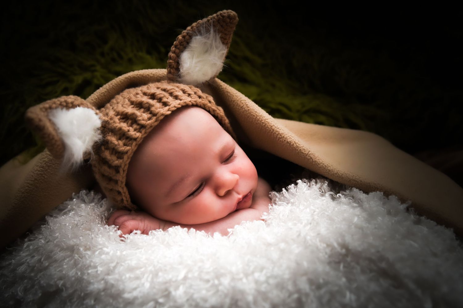 Newborn baby sleeping peacefully on soft white fur blanket wearing brown knit hat in professional studio lighting.