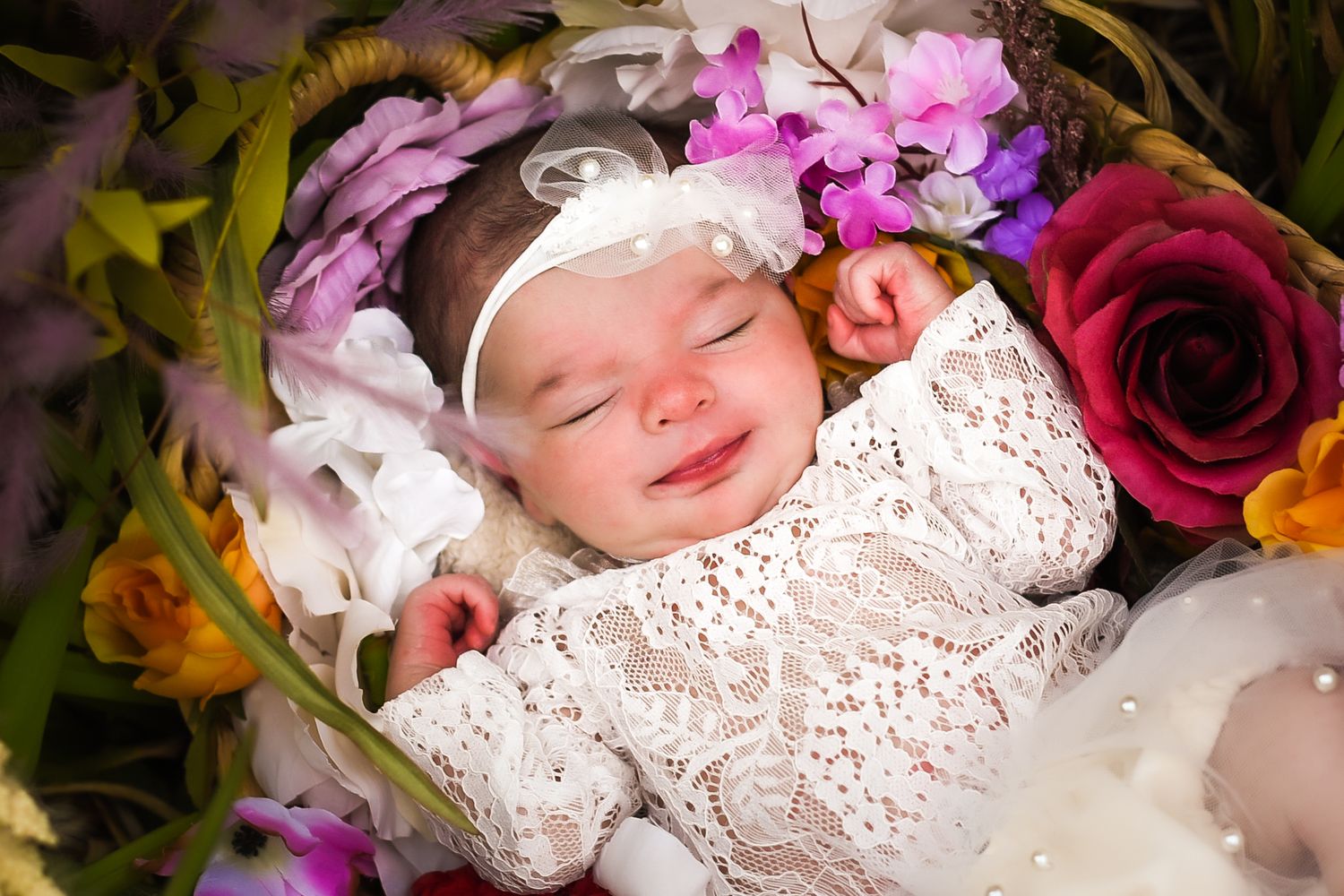A sleeping baby in white lace dress surrounded by colorful flowers and floral arrangements.