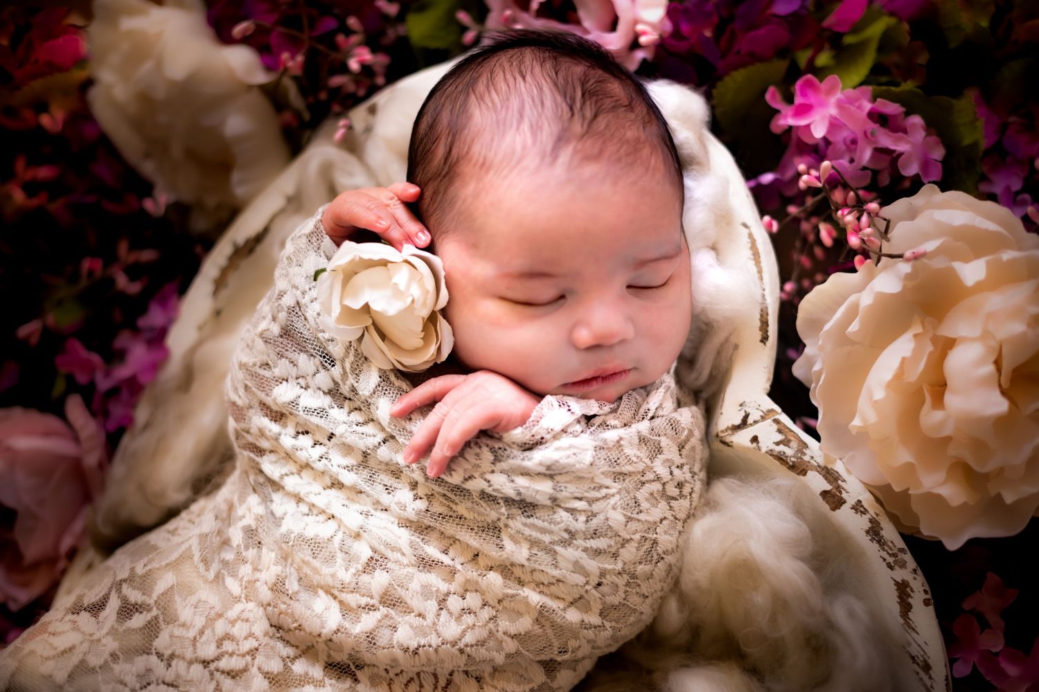 Newborn baby sleeping peacefully in a cream lace wrap surrounded by pink and peach floral decorations.