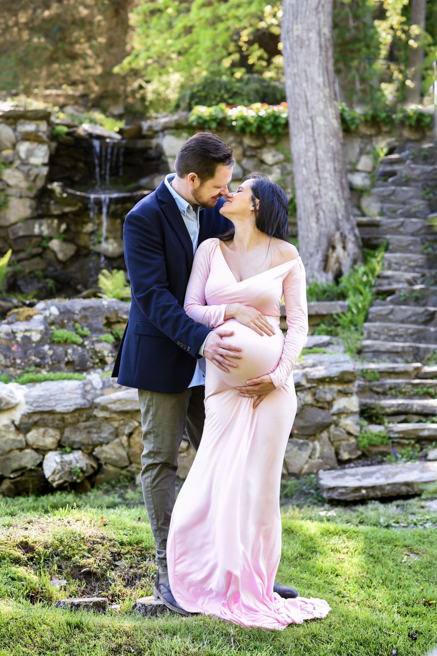 A romantic maternity photoshoot in a garden setting with stone walls and trees in the background.