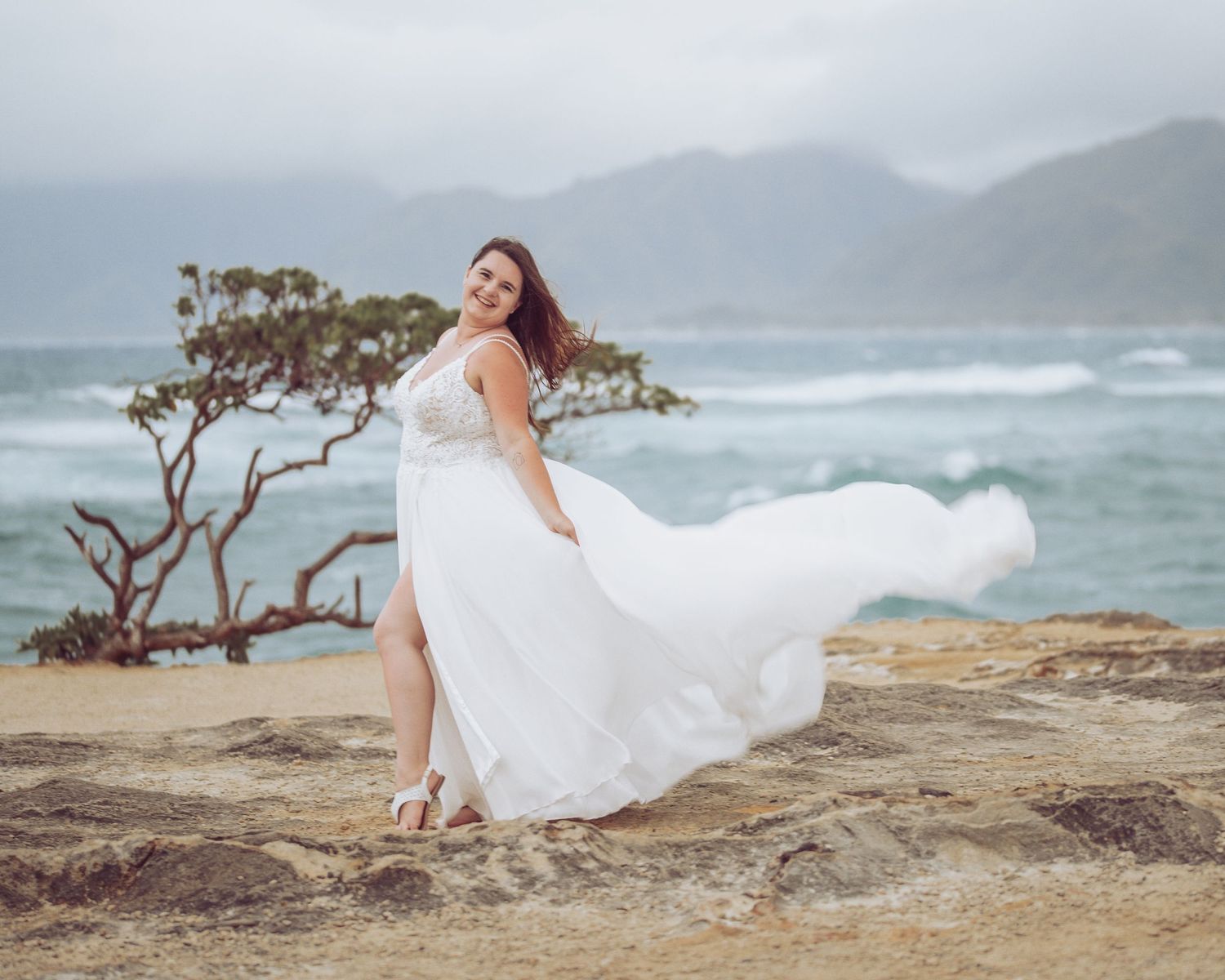 White wedding dress flows in ocean breeze on rocky coastal cliff with mountains in background.