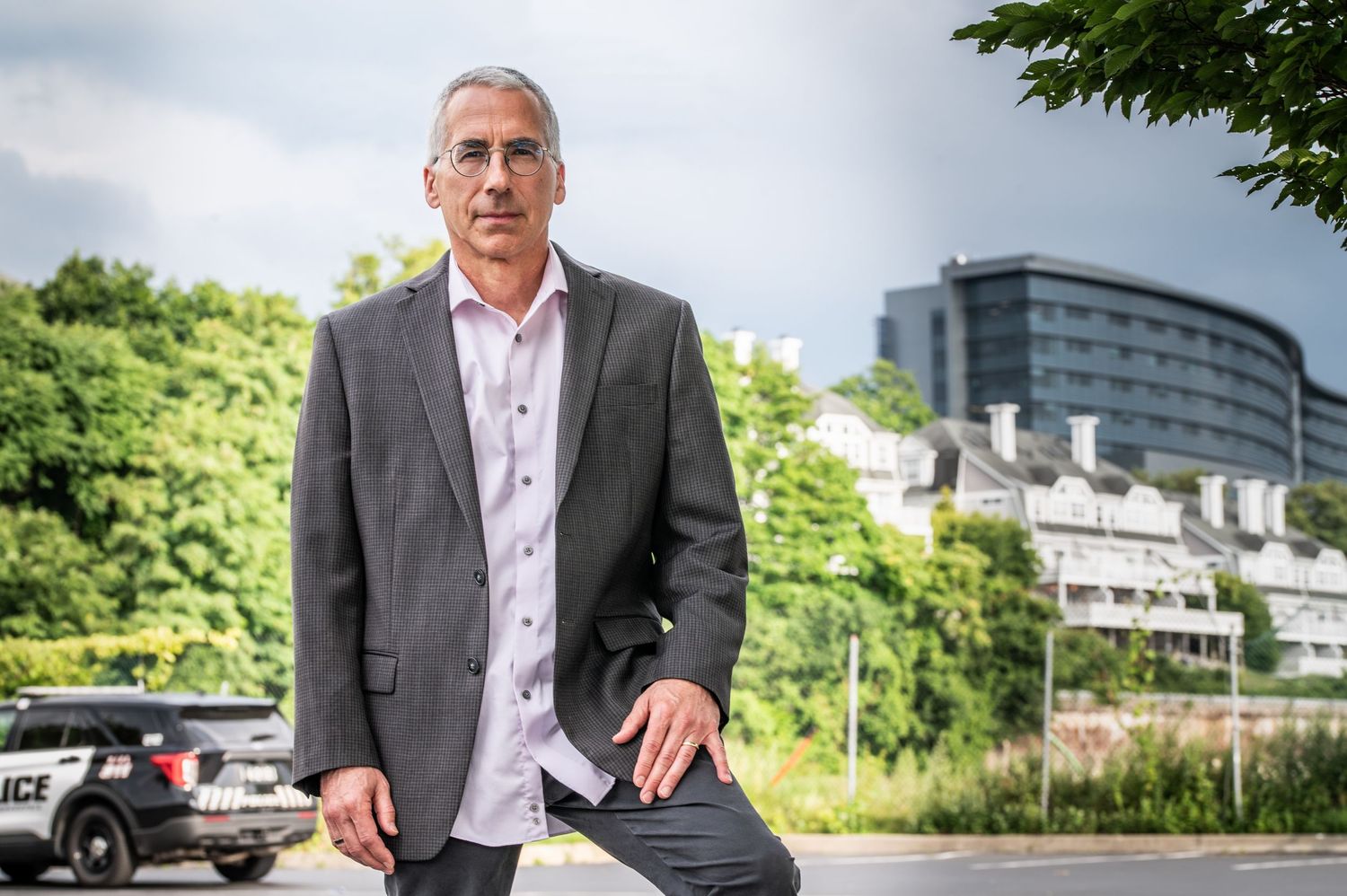 A person in a gray blazer and white shirt stands in a parking lot with office buildings and trees in the background.