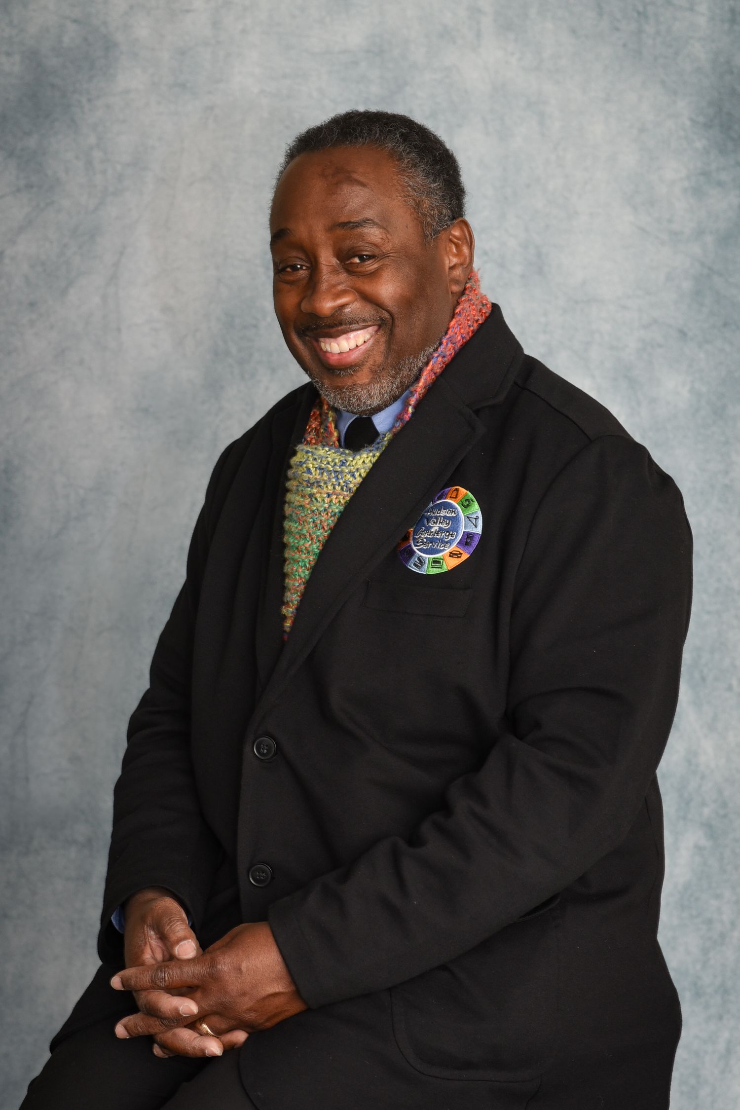 Professional portrait of a person wearing a black suit jacket with a patterned tie and pocket square against a gray background.