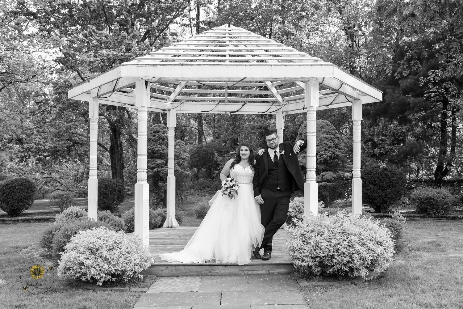 A white wooden gazebo decorated with flowers serves as a romantic wedding ceremony backdrop in a garden setting.