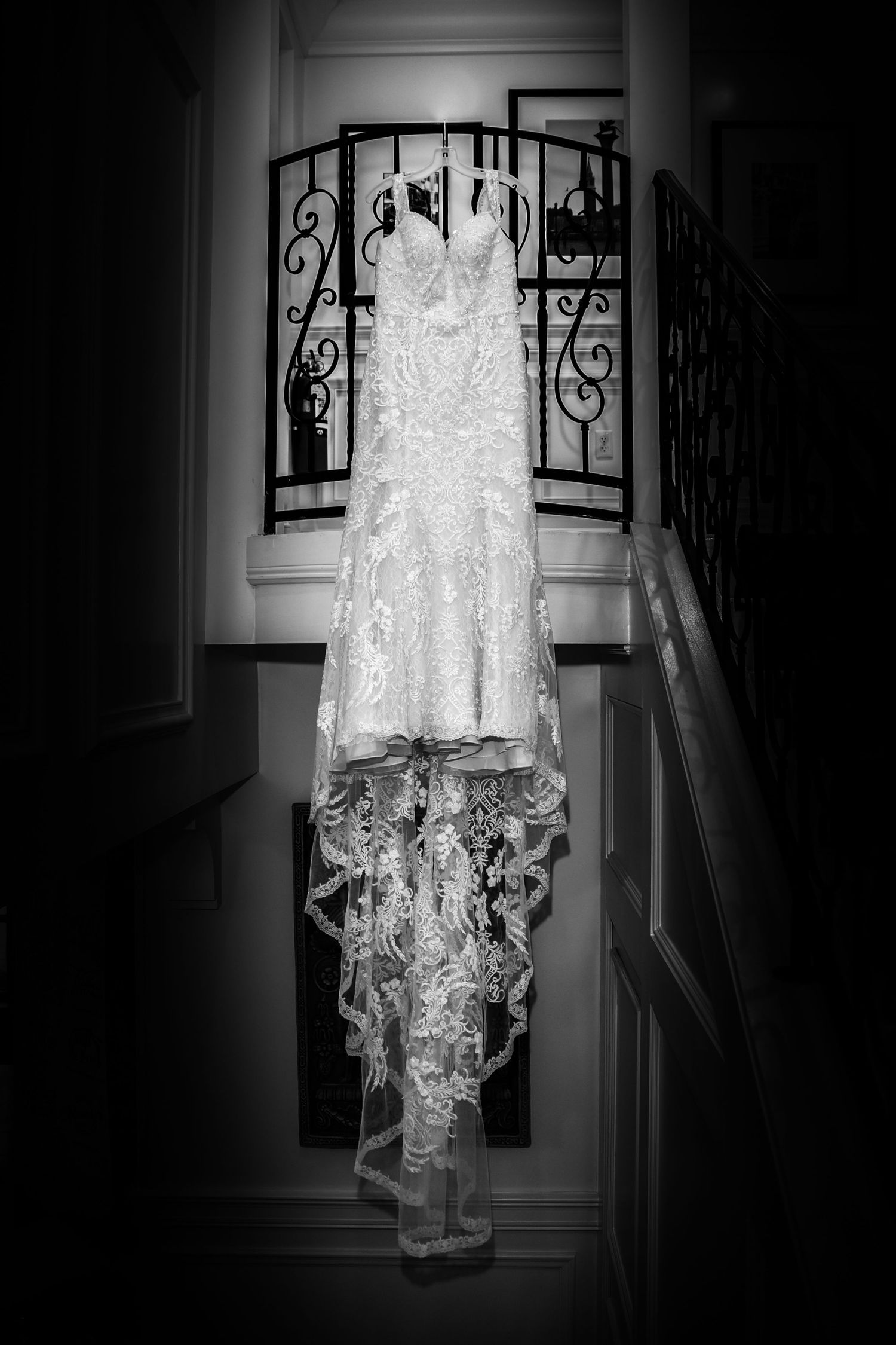 Black and white photo of a lace wedding gown suspended against ornate iron staircase railings.
