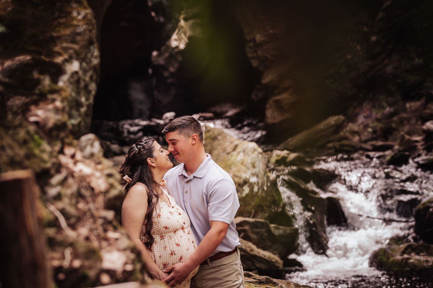 A romantic couple embraces near a cascading waterfall in a scenic forest setting during an engagement photoshoot.