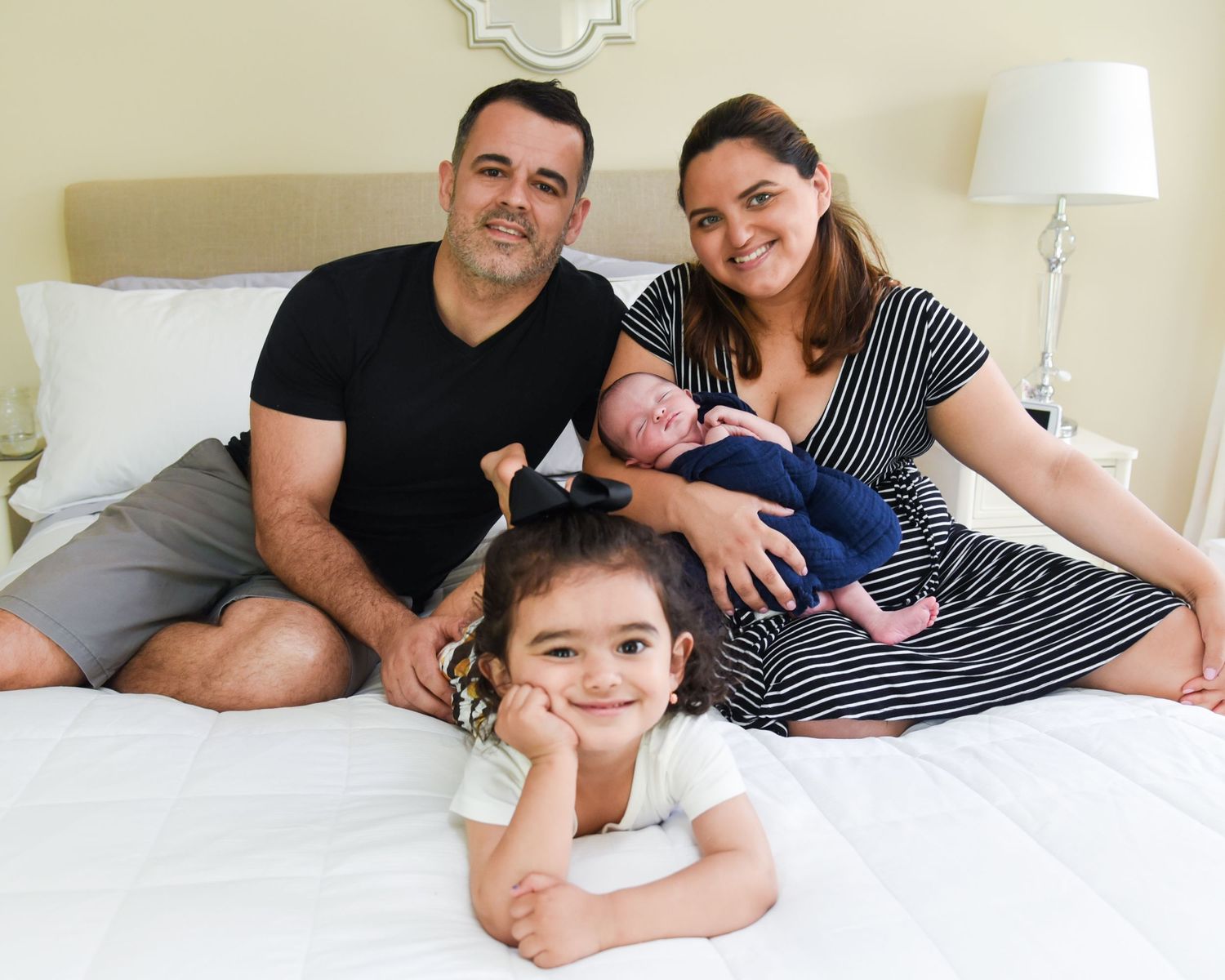 A family lies together on a white bed wearing casual clothes in a bright bedroom setting.