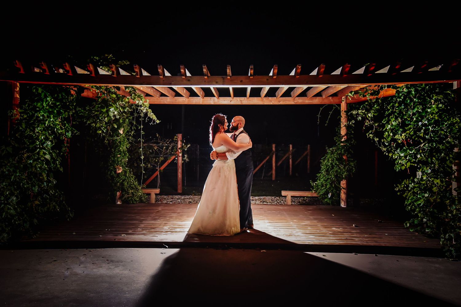 A couple shares a romantic dance under a wooden pergola surrounded by lush greenery at sunset.