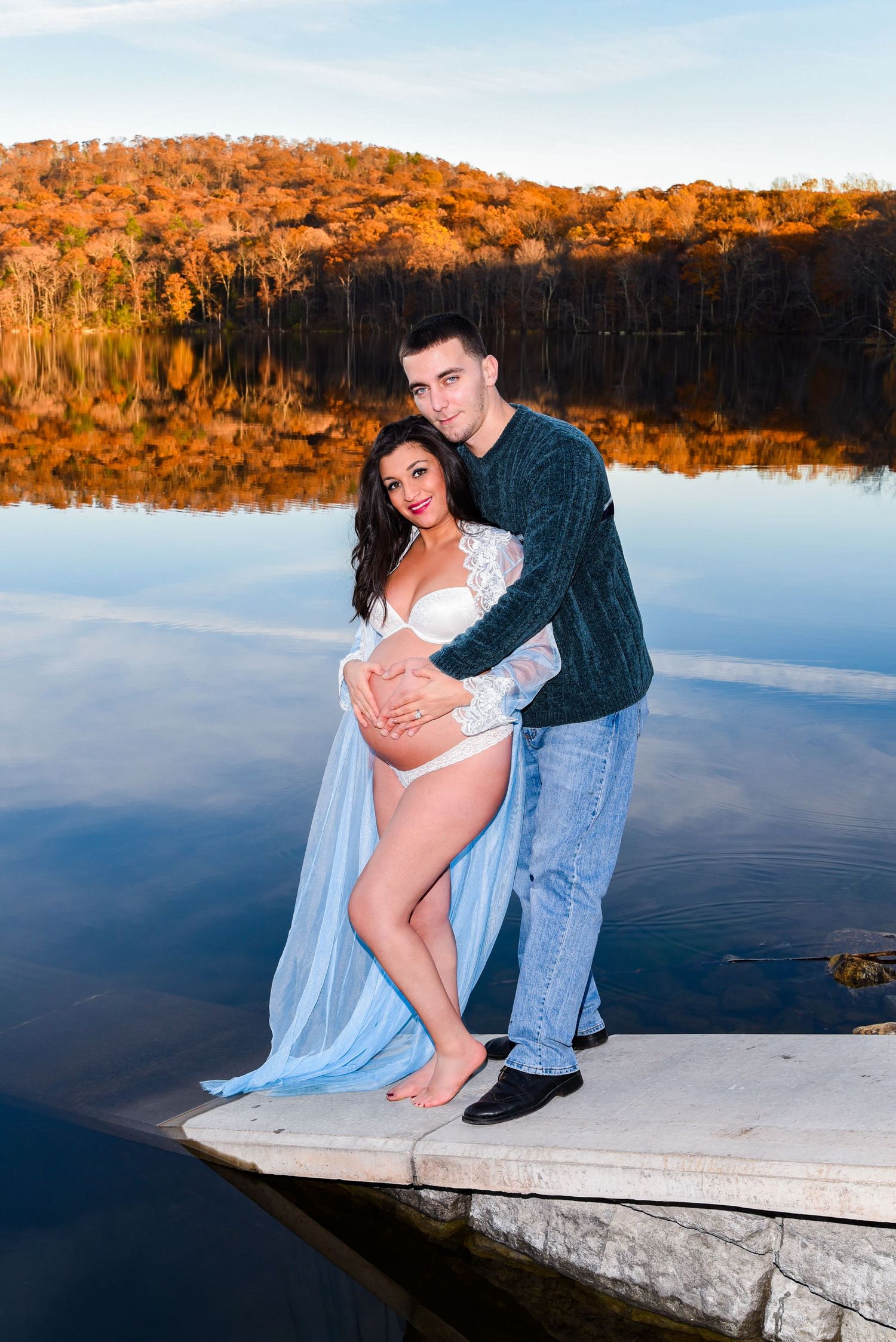 A couple poses romantically on a wooden dock by a serene lake during golden hour with autumn foliage reflecting in the water.