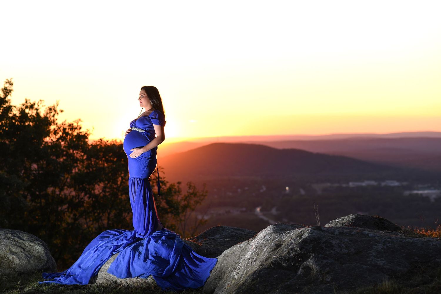 A person in a flowing royal blue dress stands on a rocky overlook at sunset against a mountain landscape.