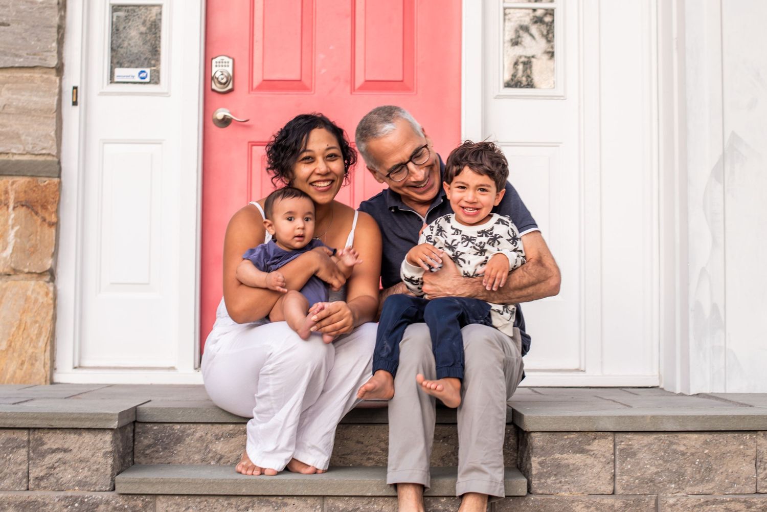 A family sits together smiling on stone steps in front of a bright coral-colored door.