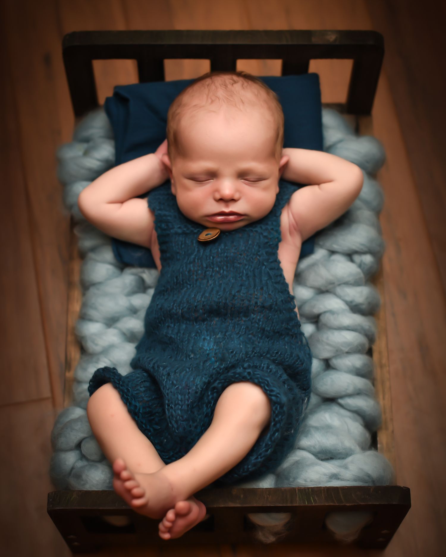 Sleeping baby wearing knitted teal romper lies on soft blue bedding in wooden crib with arms behind head.