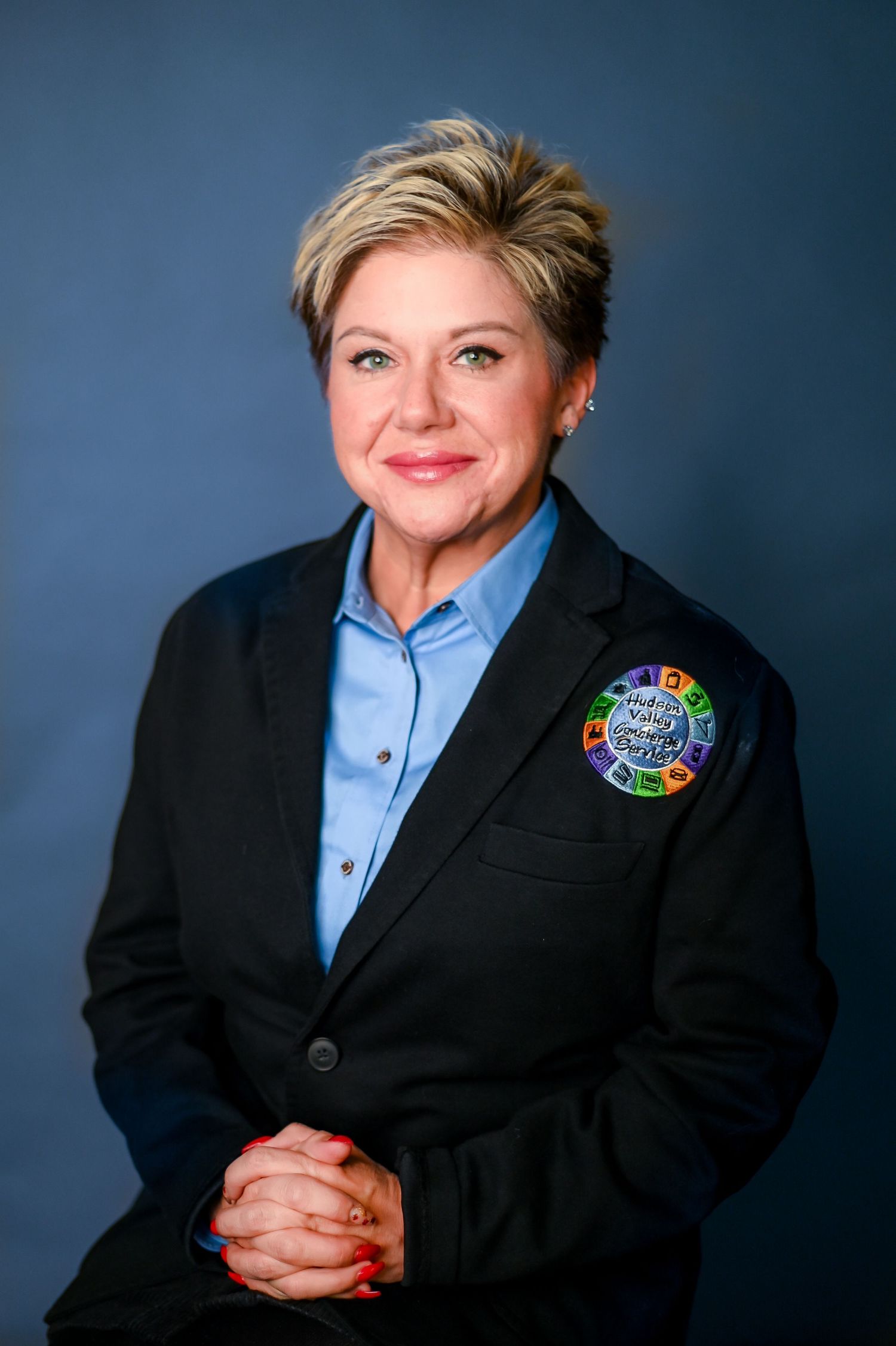 Professional portrait of someone wearing a blue button-down shirt, black blazer and circular rainbow-colored pin against blue backdrop.