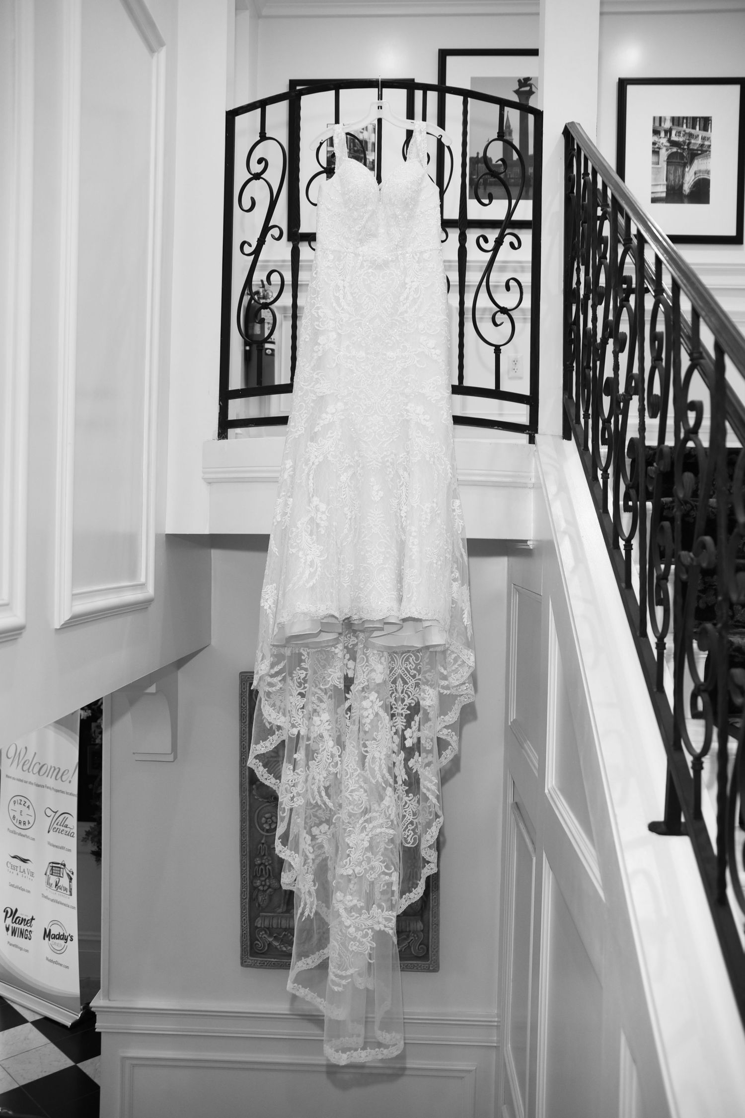 Elegant lace wedding dress hanging on ornate black iron railing in a white hallway.