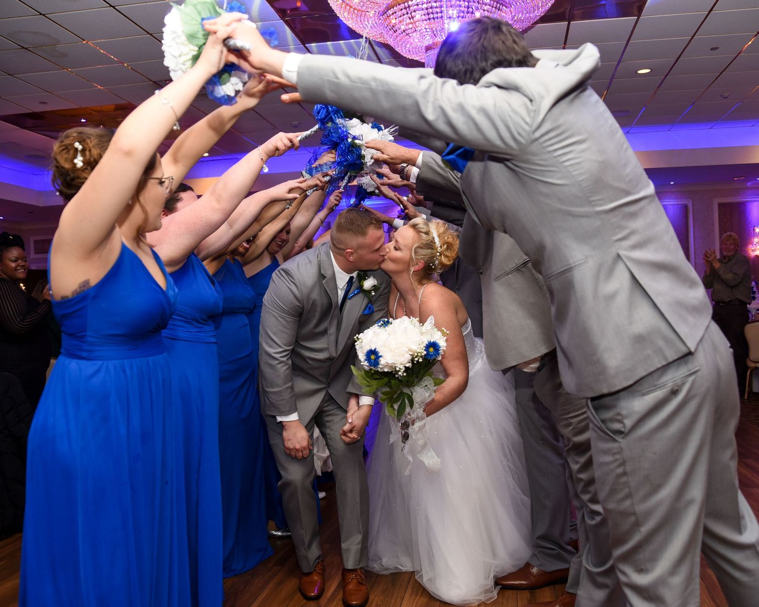 Wedding party dips the couple during a dance with bridesmaids in blue dresses and groomsmen in gray suits.