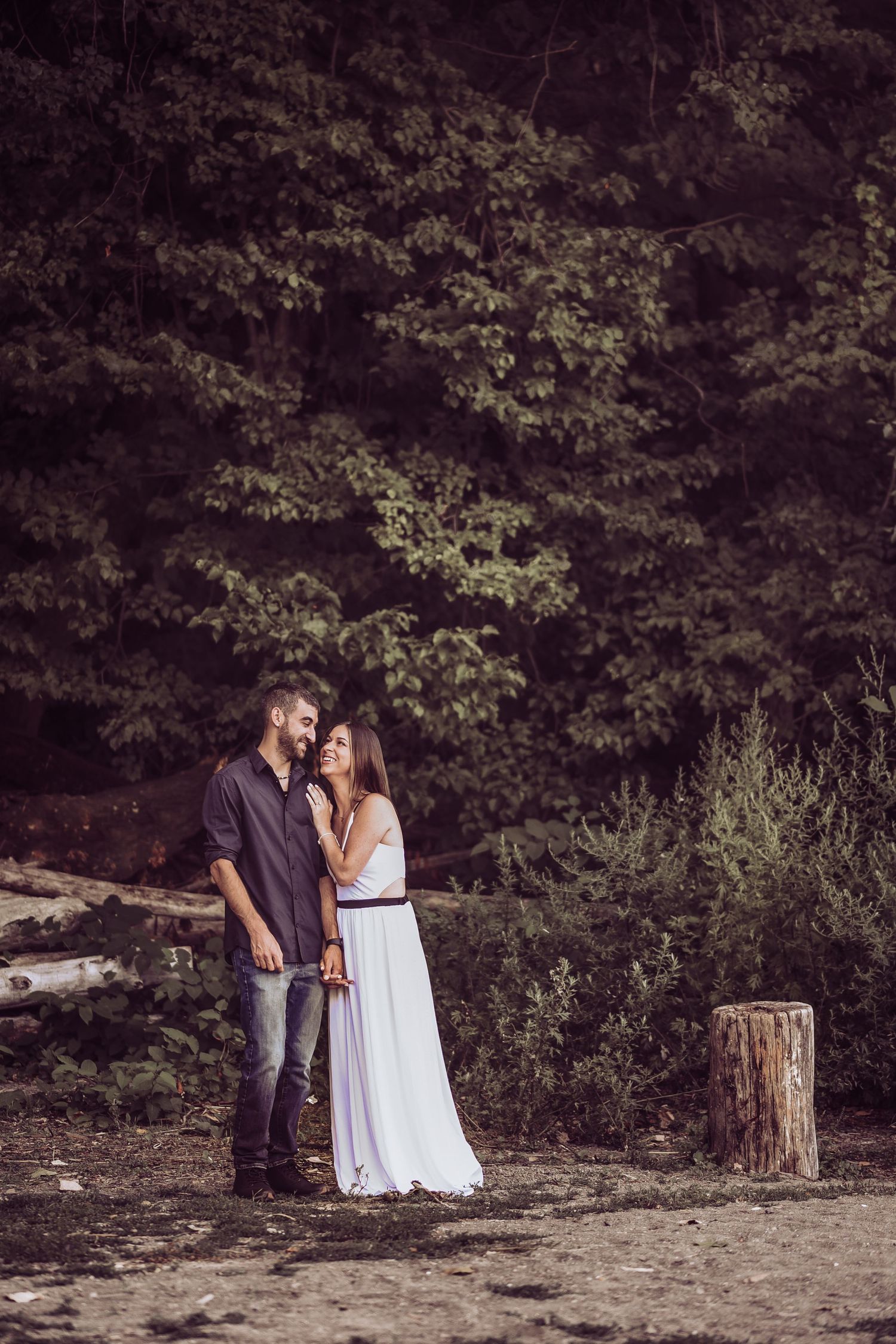 Romantic couple in white dress and dark attire embracing in a wooded outdoor setting.