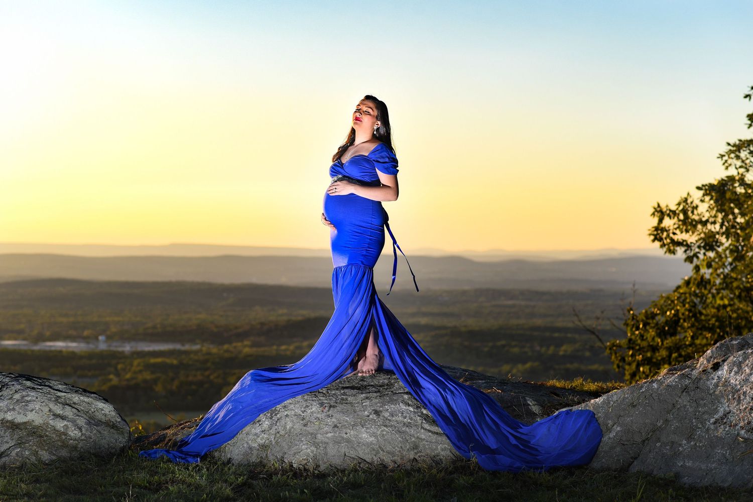 A woman in a flowing blue gown poses dramatically on a rocky overlook during golden hour.