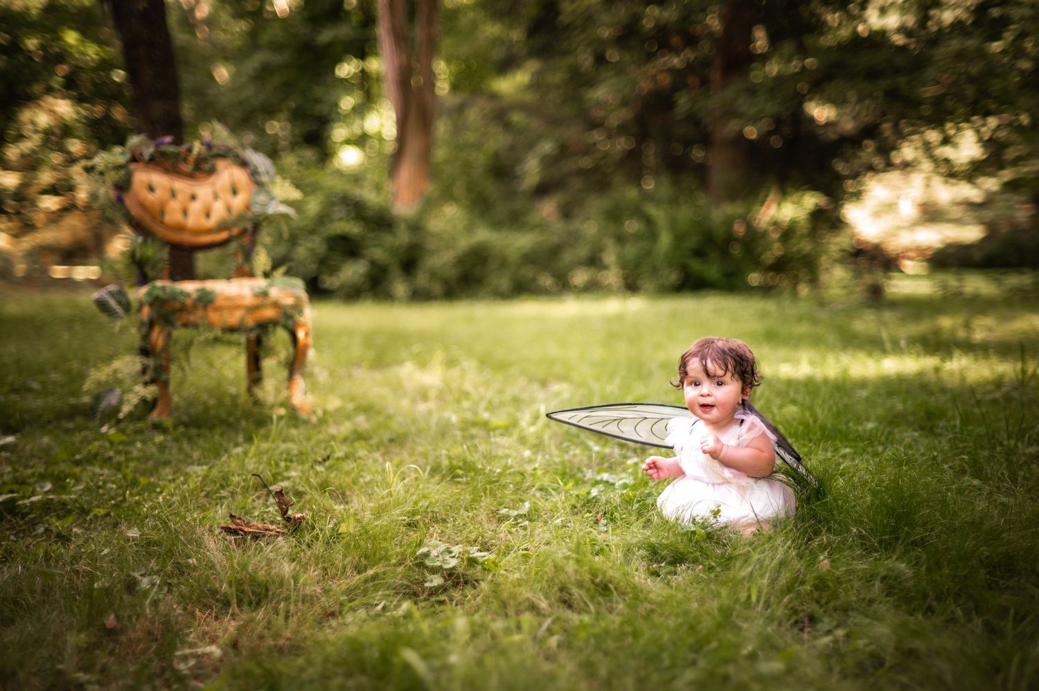 Little one in white dress sitting on grass near vintage chair in dreamy garden setting.