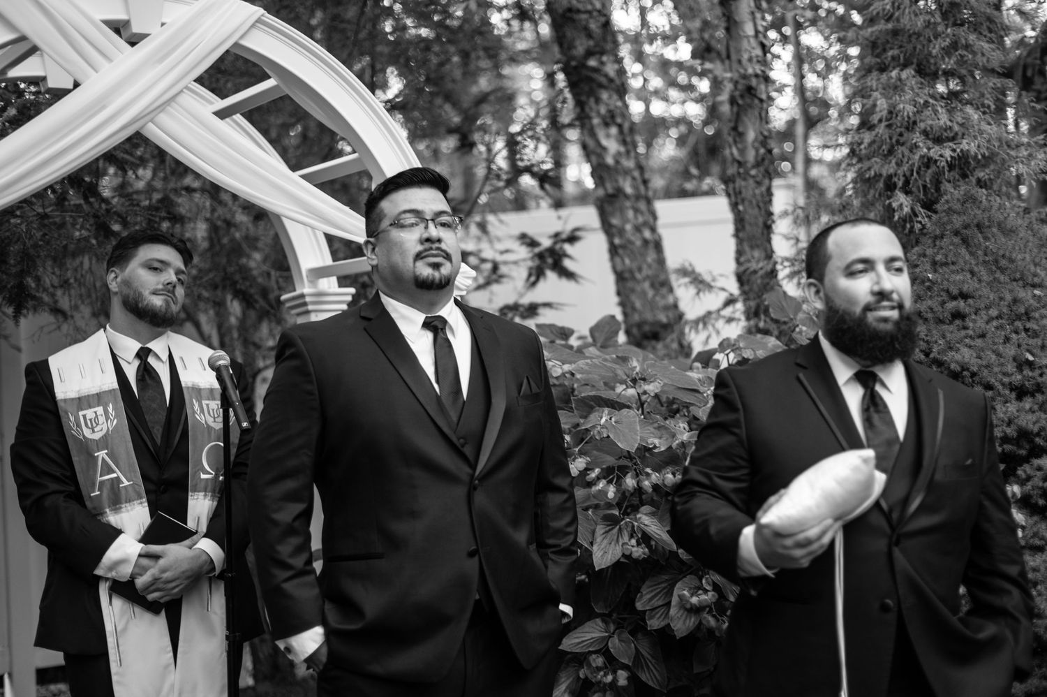 Three groomsmen in dark suits stand during an outdoor wedding ceremony.