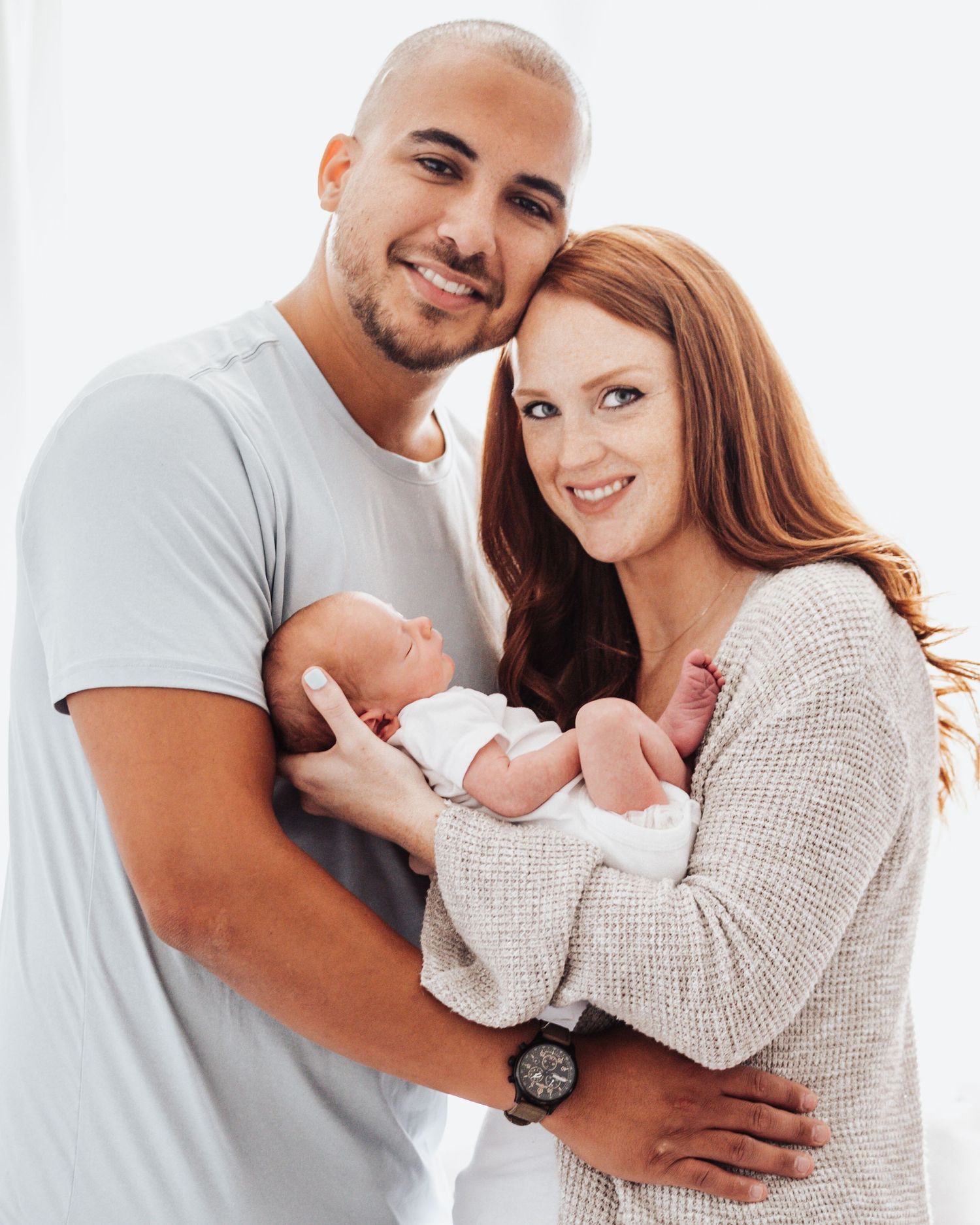 Happy couple in white and grey clothing embracing while holding their newborn baby.
