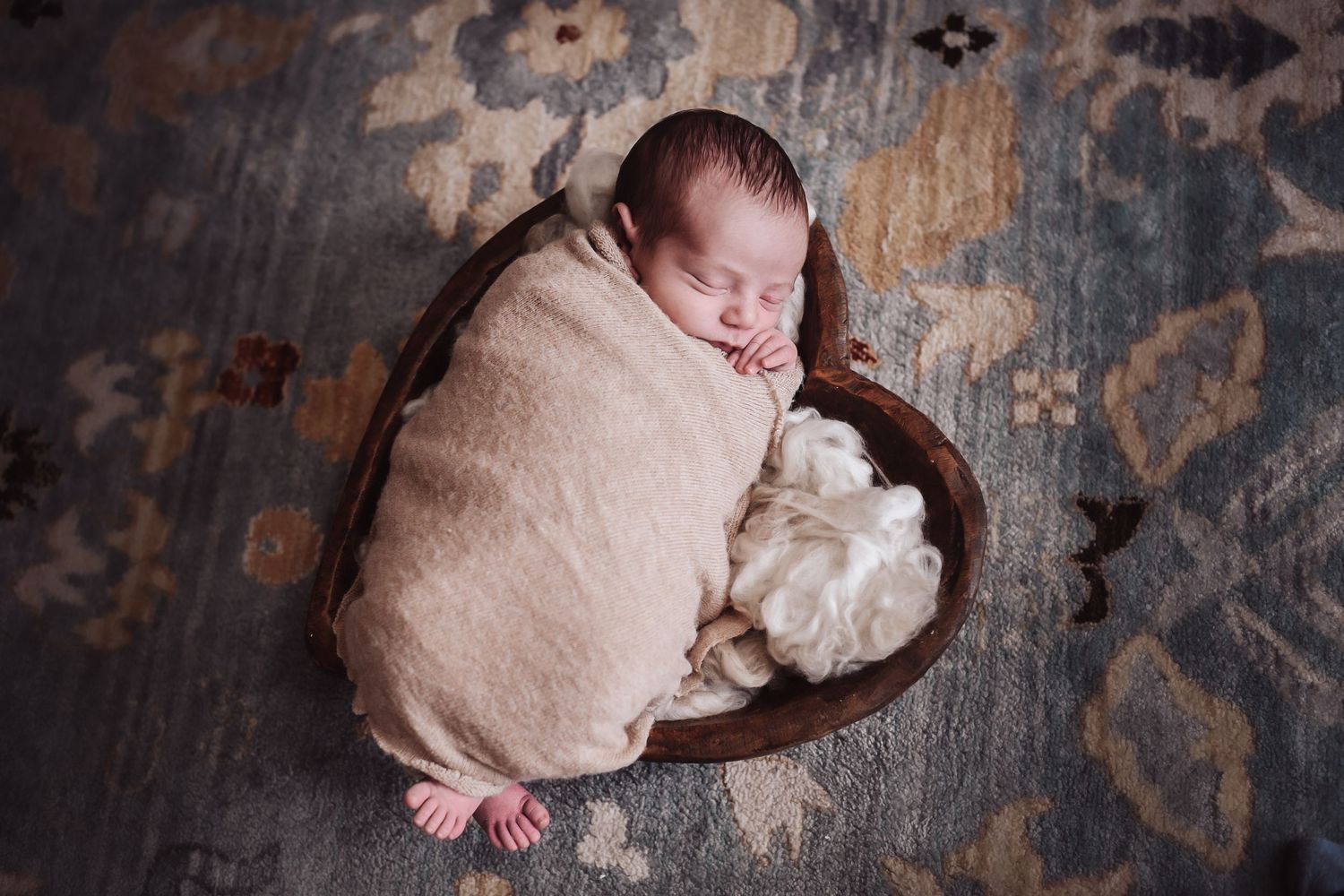 A sleeping newborn baby wrapped in a soft pink swaddle blanket rests on a dark wooden surface.