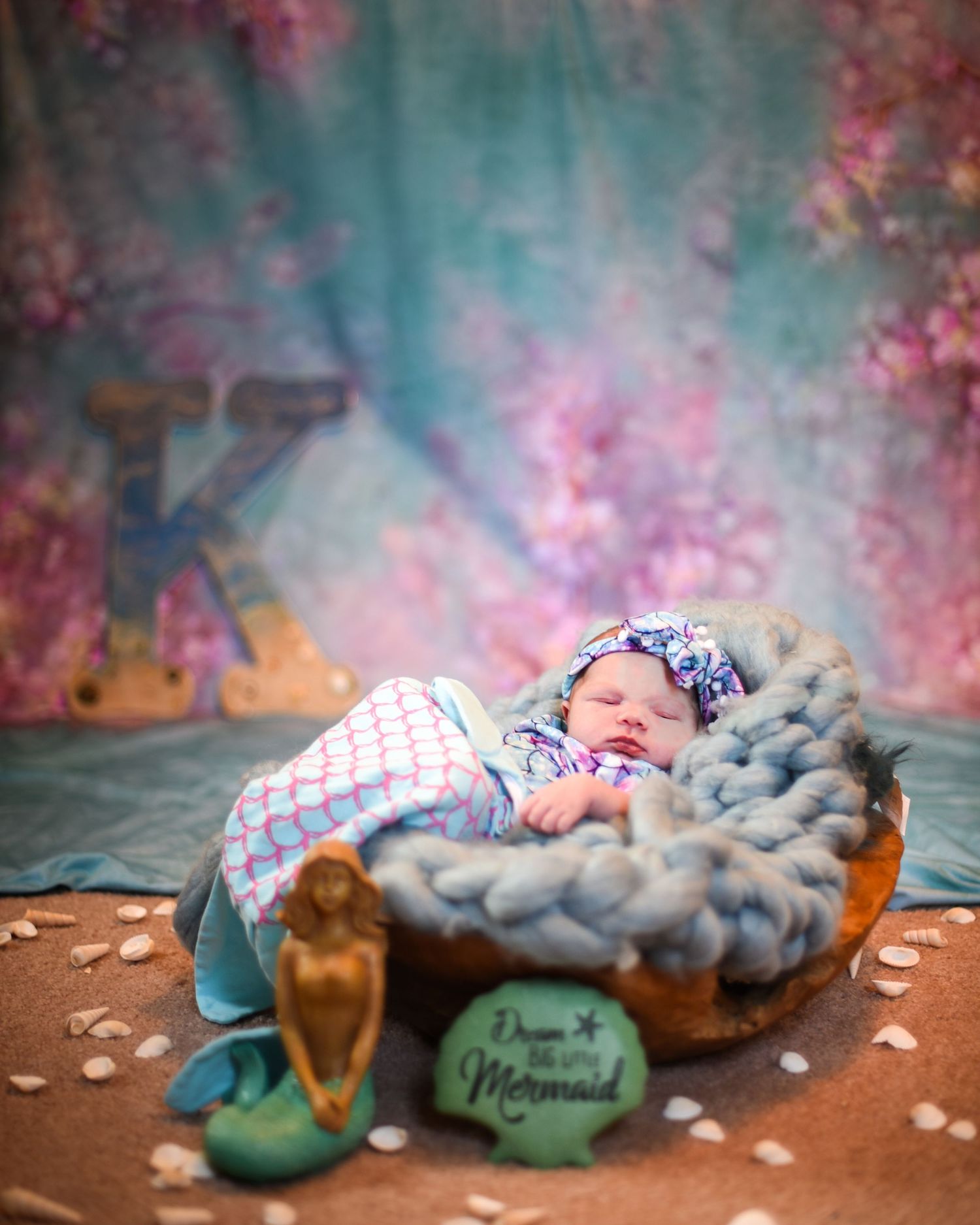 A sleeping baby wrapped in a checkered blanket lies on a wooden surface with dreamy pink and teal bokeh background effects.