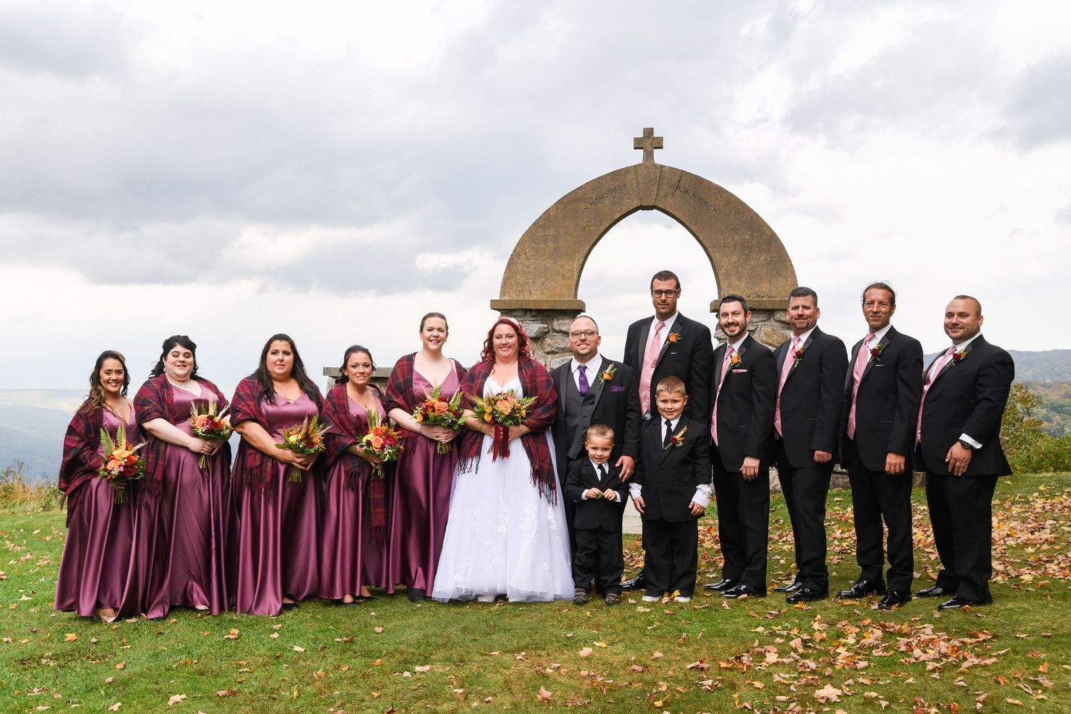 Wedding party in burgundy and black attire poses outdoors near stone monument.