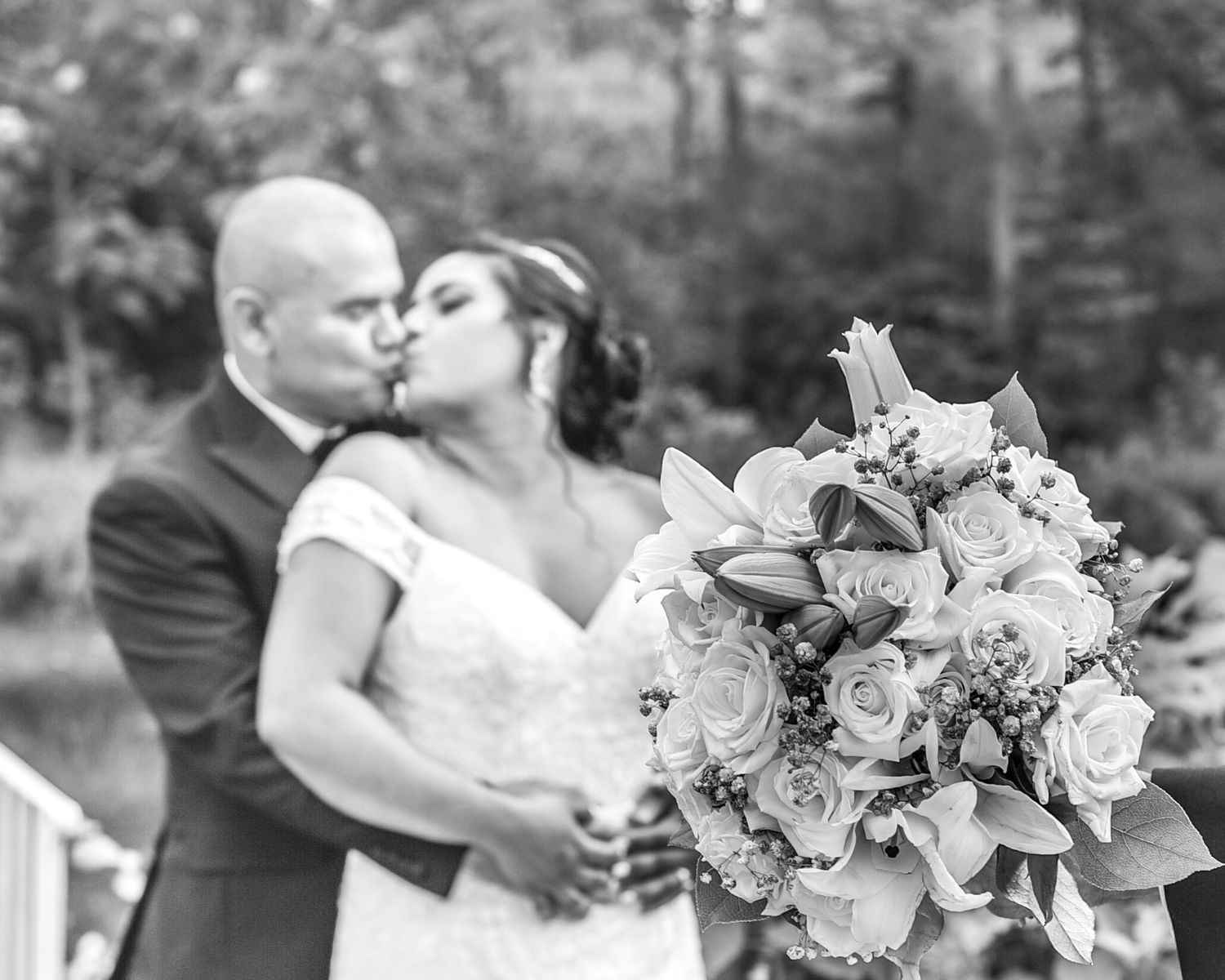 Black and white photo of couple kissing with bridal bouquet of roses in foreground.
