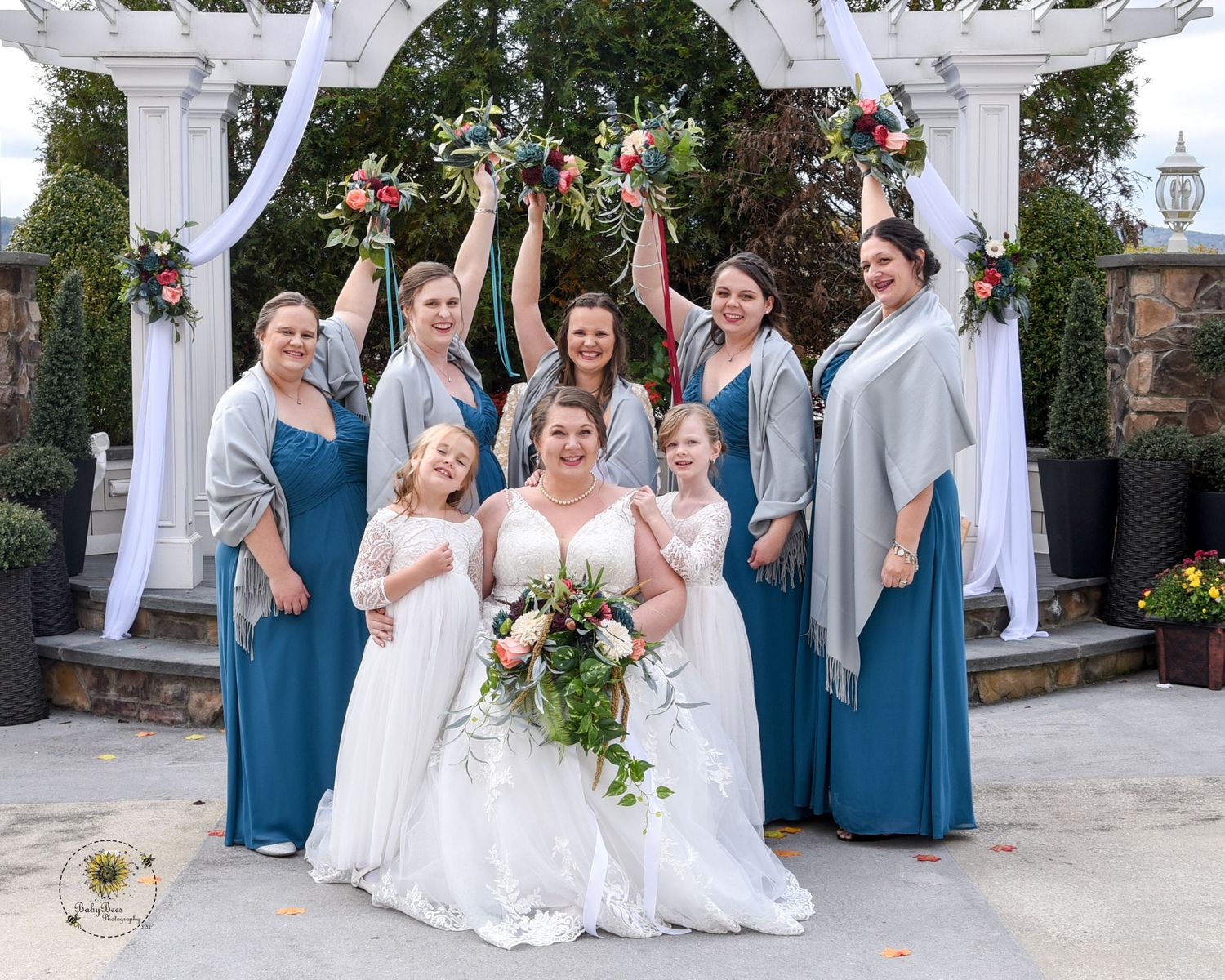Bridal party in blue dresses and white gowns poses under garden archway.
