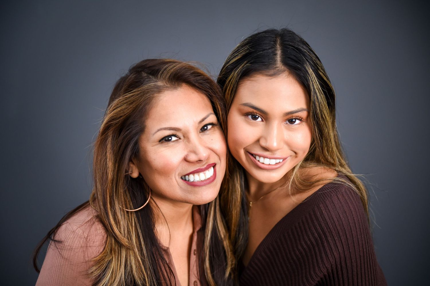Two friends smiling and embracing while wearing brown sweaters against a dark background.