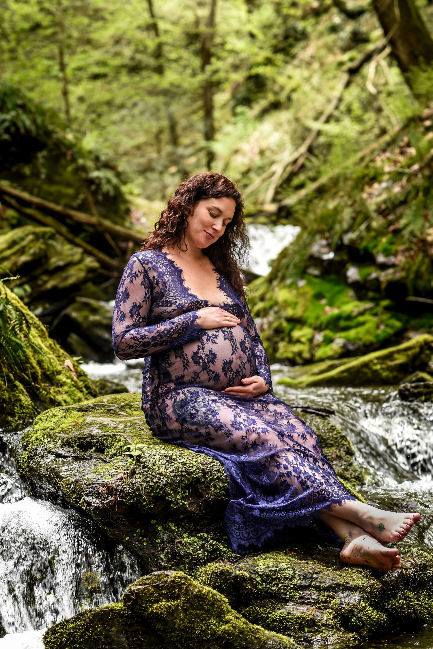 A pregnant woman in a blue lace dress poses gracefully by a forest stream and mossy rocks in a nature maternity photoshoot.