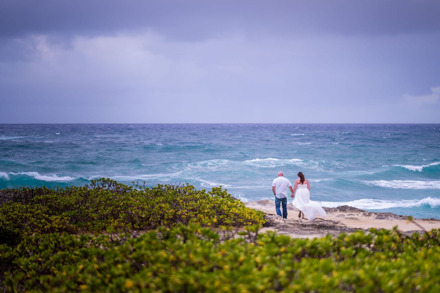 Two figures stand on a scenic coastal cliff overlooking turquoise waters with green vegetation in the foreground.