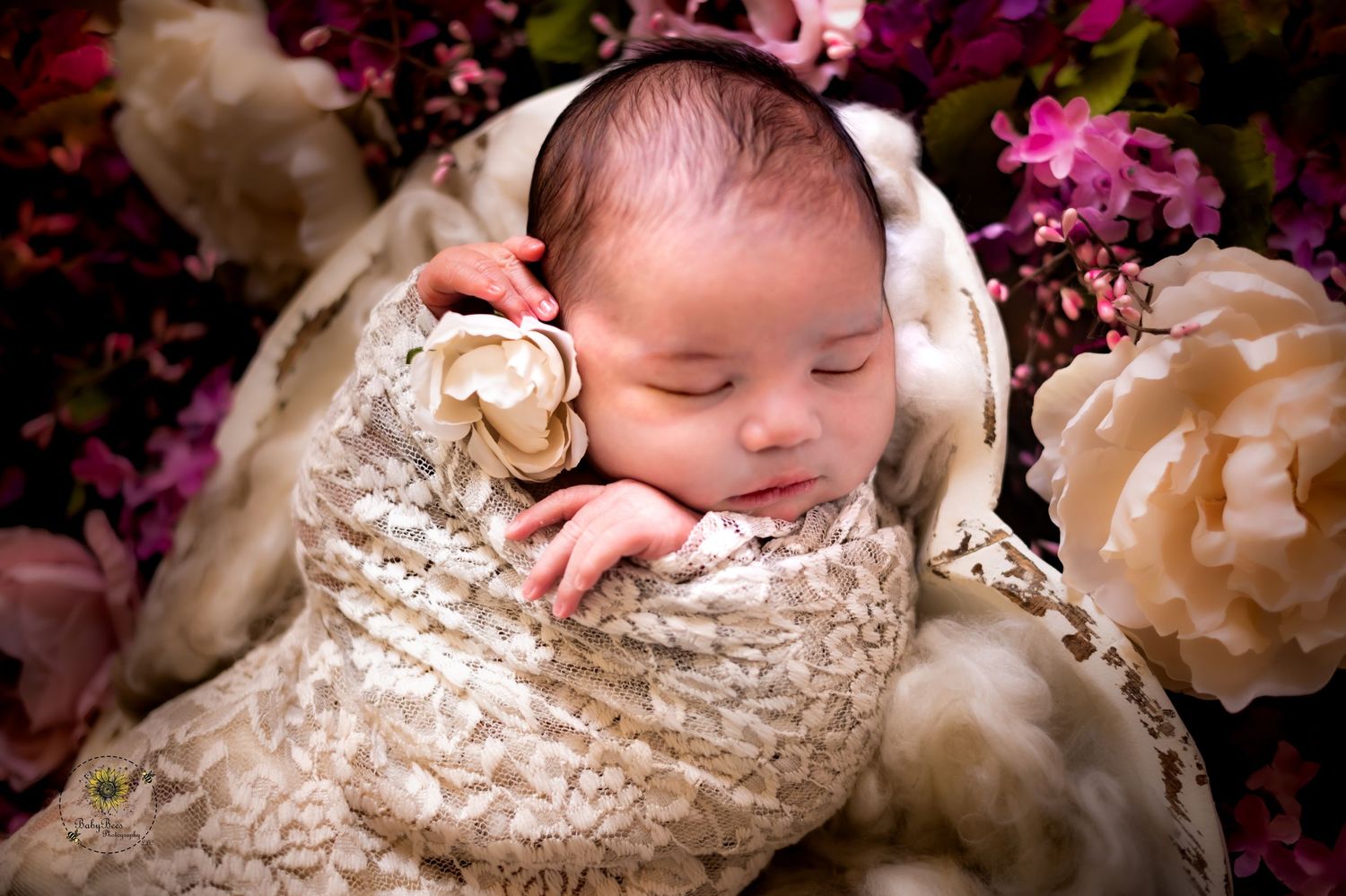A newborn baby sleeps peacefully in a cream lace wrap surrounded by pink and burgundy floral decorations.