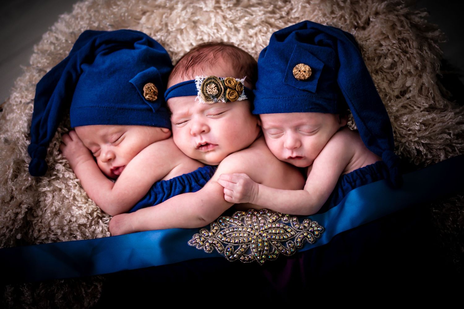 Three sleeping newborns wearing matching blue knit caps lie side by side on a textured blanket.