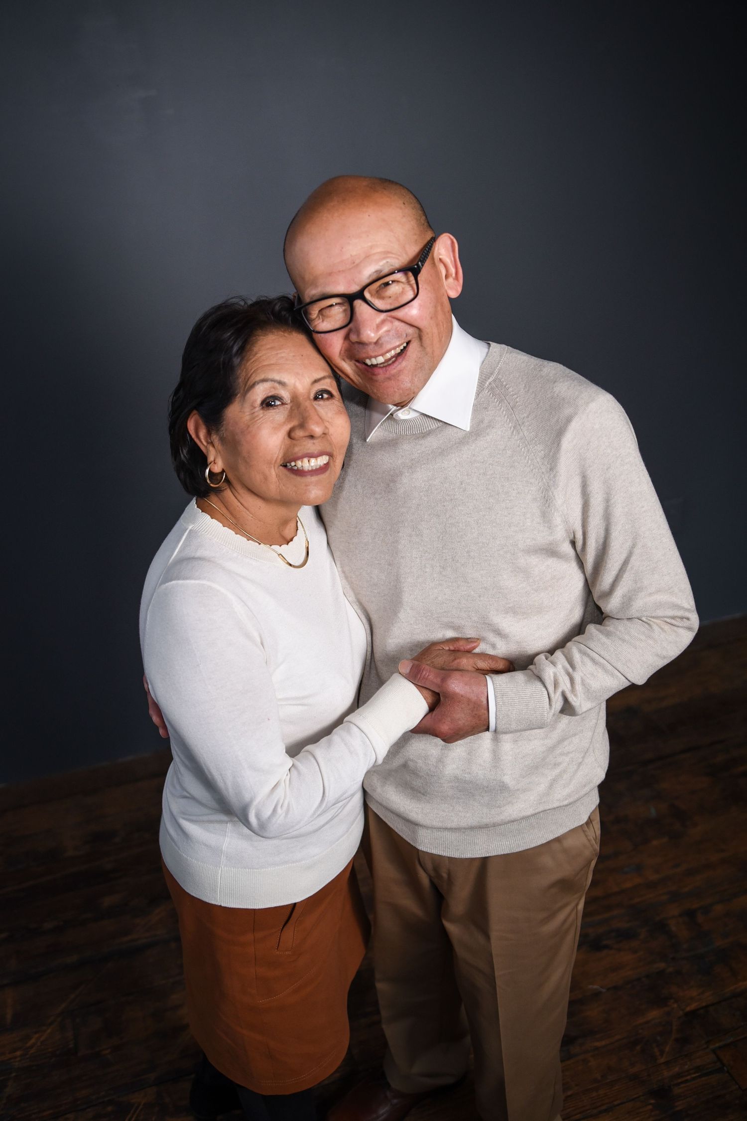Happy senior couple embracing and smiling together while wearing matching white sweaters against dark background.