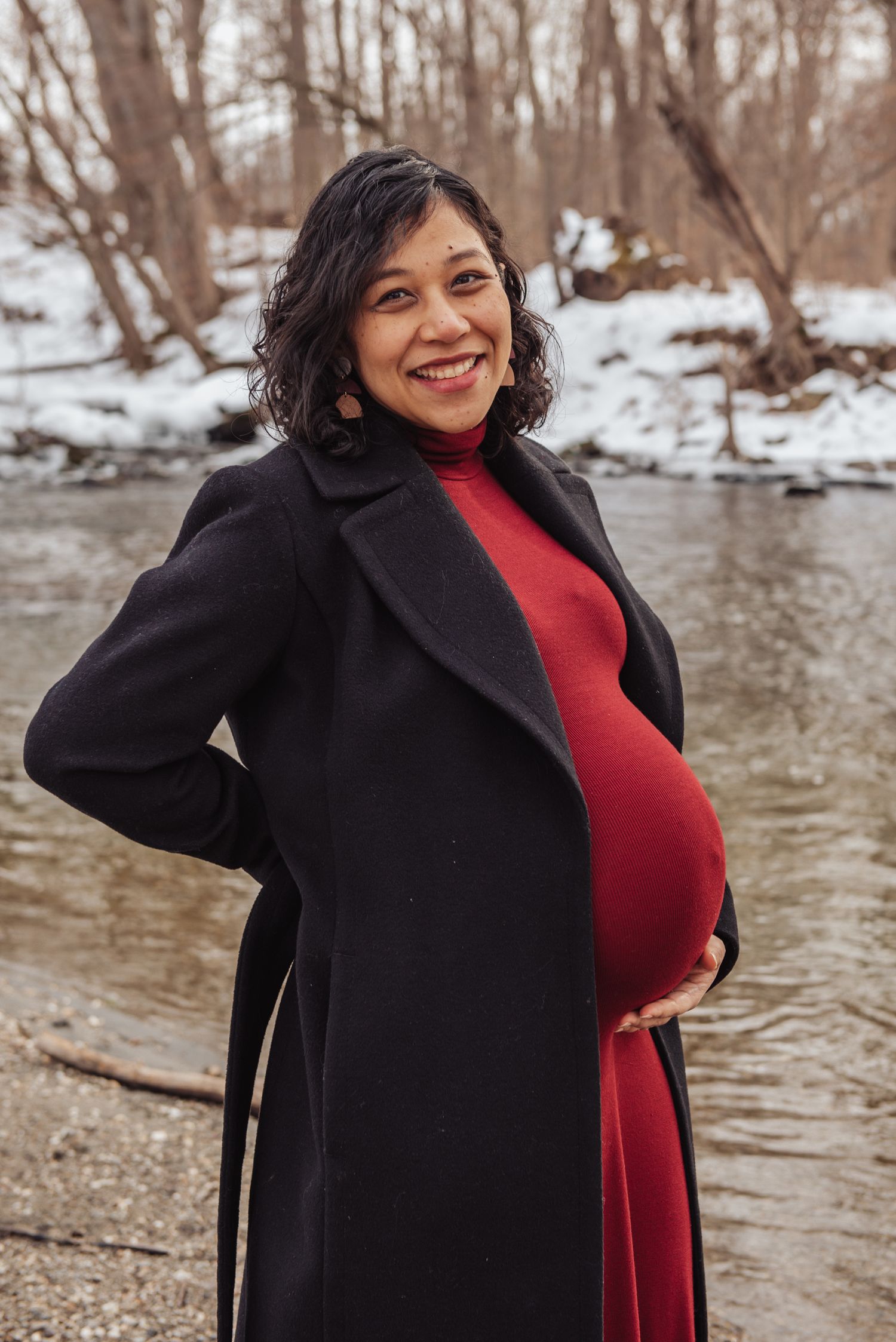 A person in a black coat and red sweater poses by a snowy riverbank in winter.