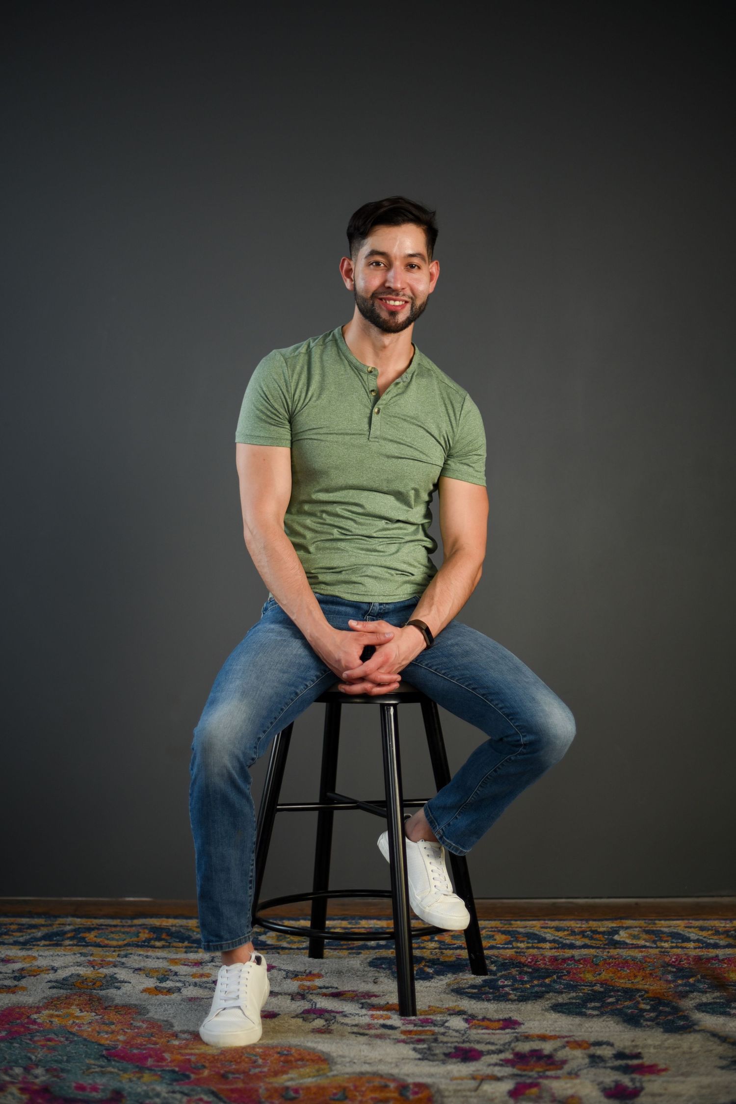 Person in green henley shirt and jeans sits casually on black stool against dark backdrop with colorful rug below.