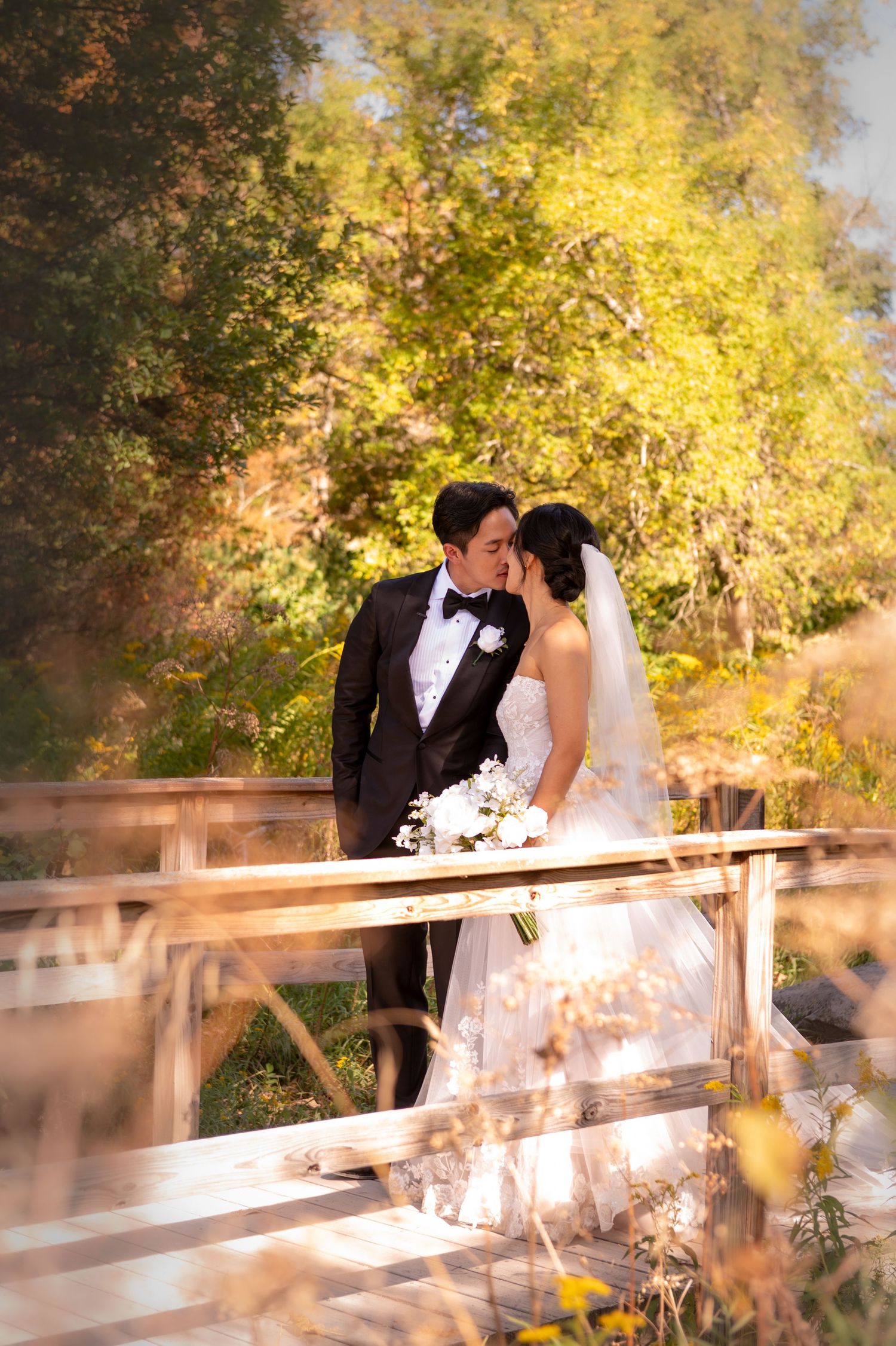 A romantic wedding couple shares tender moments on a wooden bridge surrounded by golden autumn foliage at sunset.