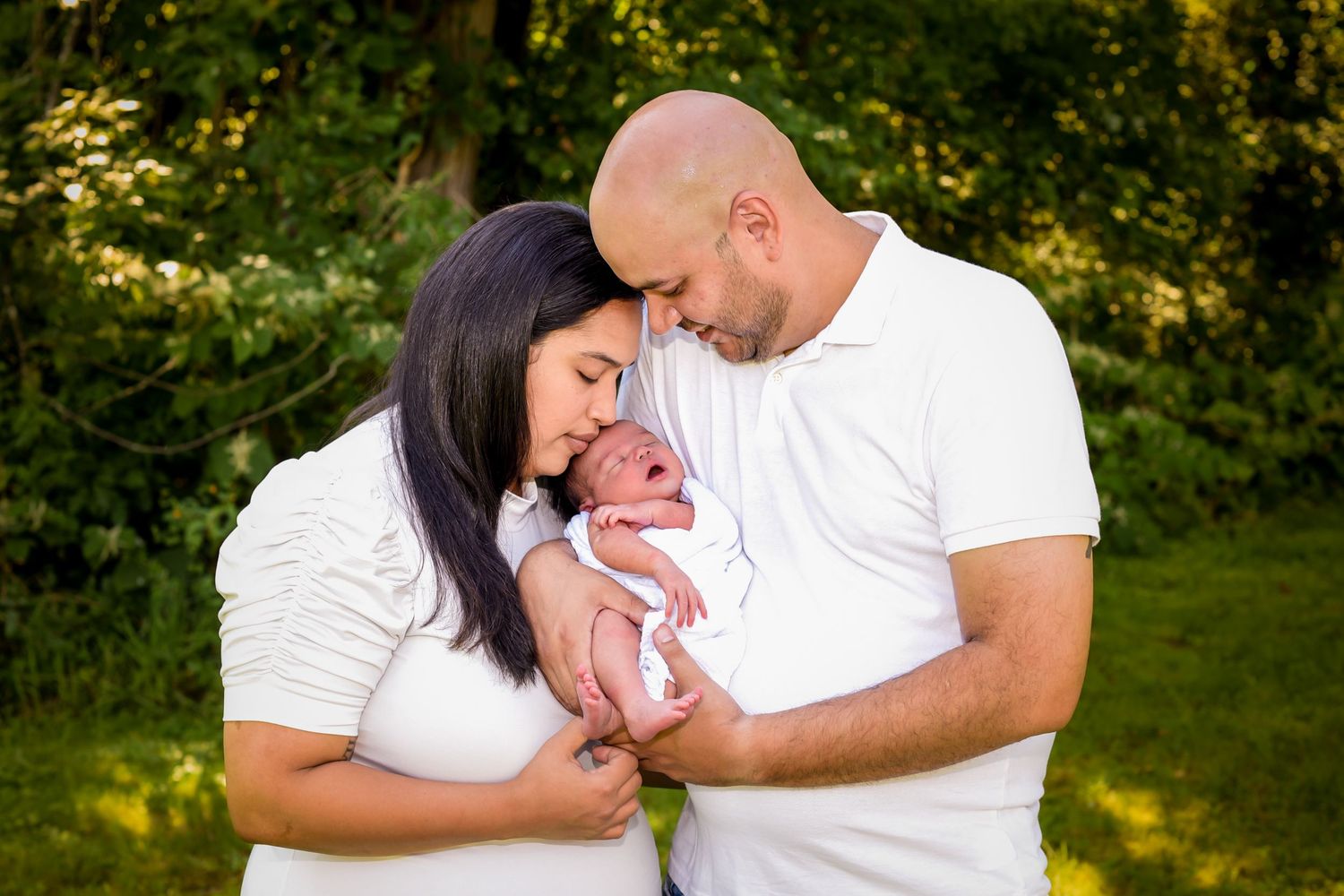 A couple in matching white shirts shares a tender moment with their newborn baby outdoors in nature.