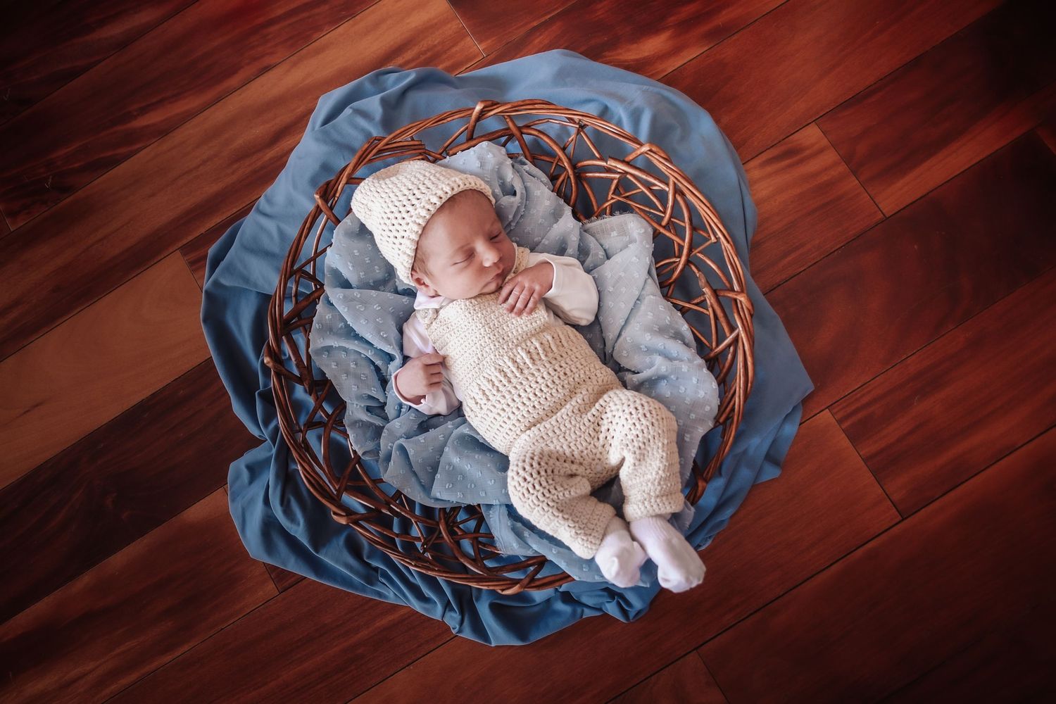 Sleeping newborn baby in white outfit nestled in blue fabric and woven wreath on wooden floor.