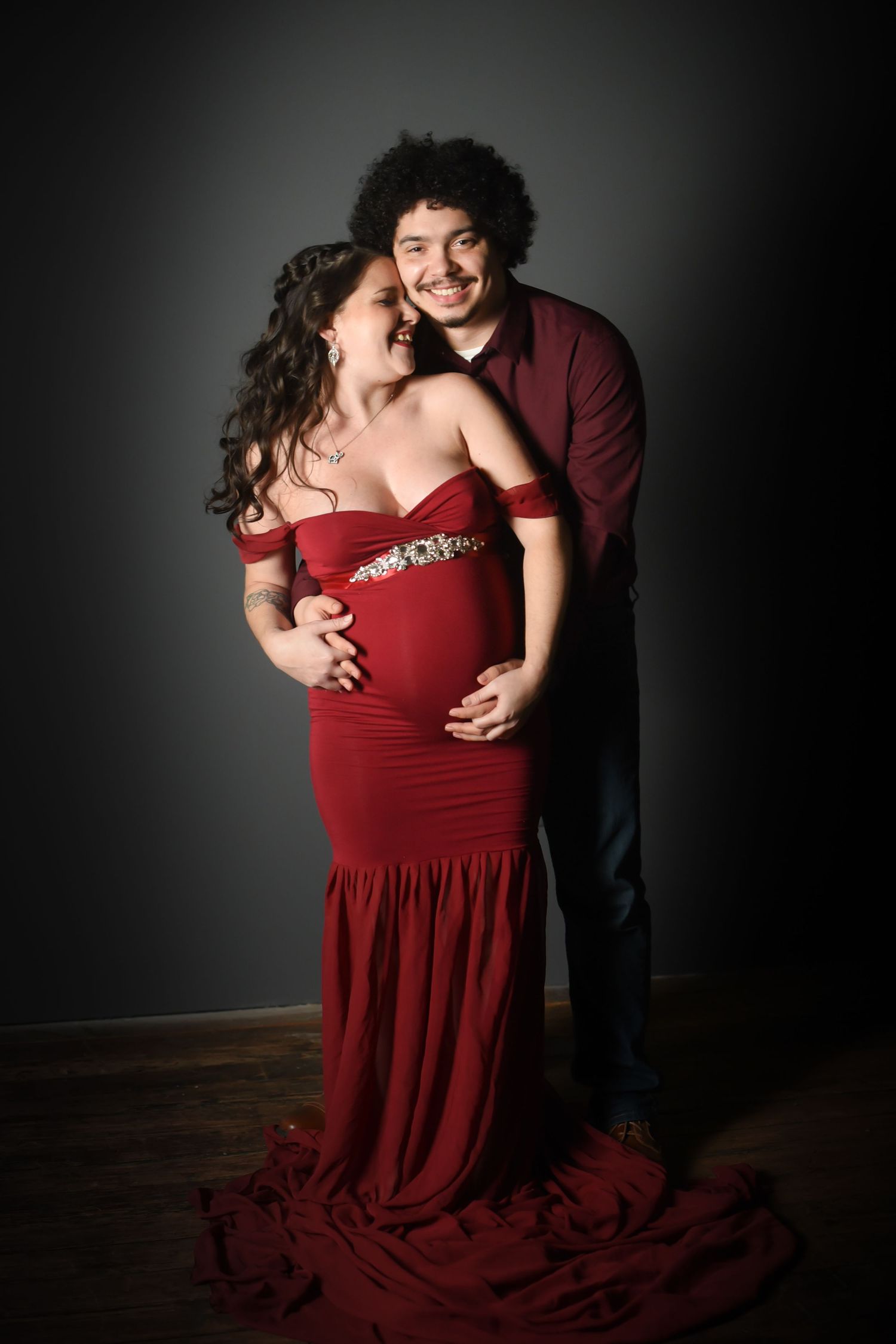 A couple embraces intimately in a romantic photoshoot, with the woman wearing a flowing red dress against a dark background.