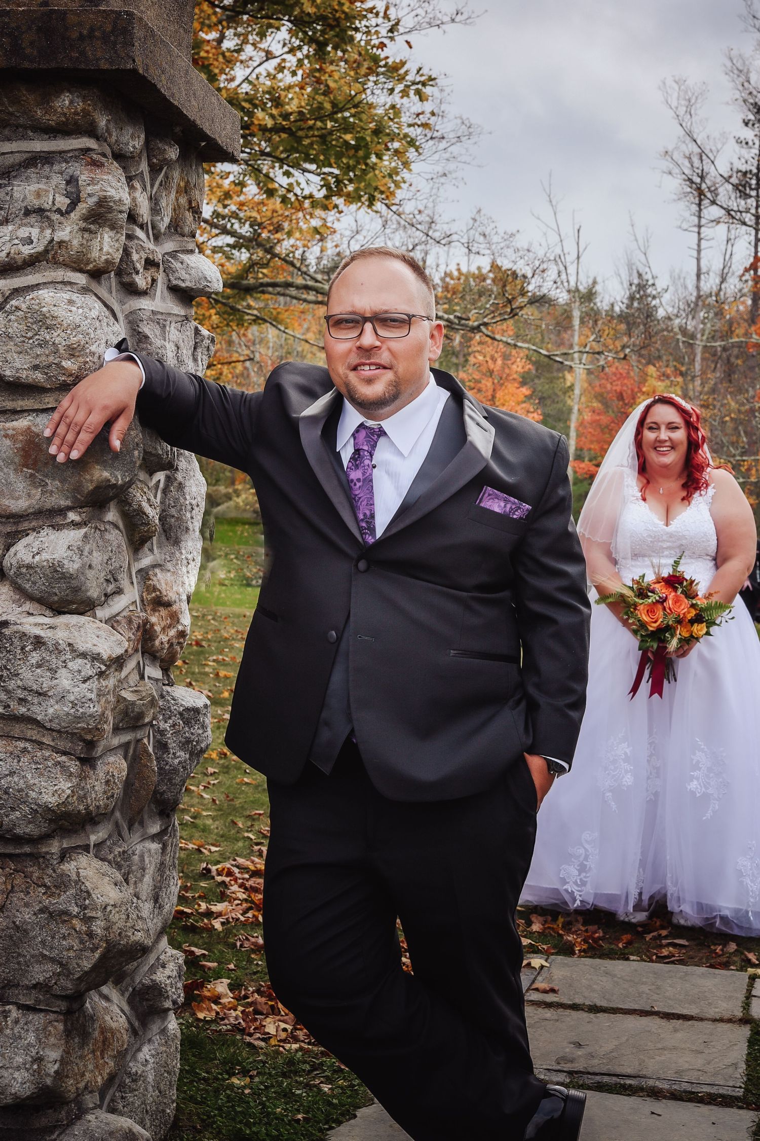Wedding couple poses by stone wall during autumn with colorful fall foliage in background.