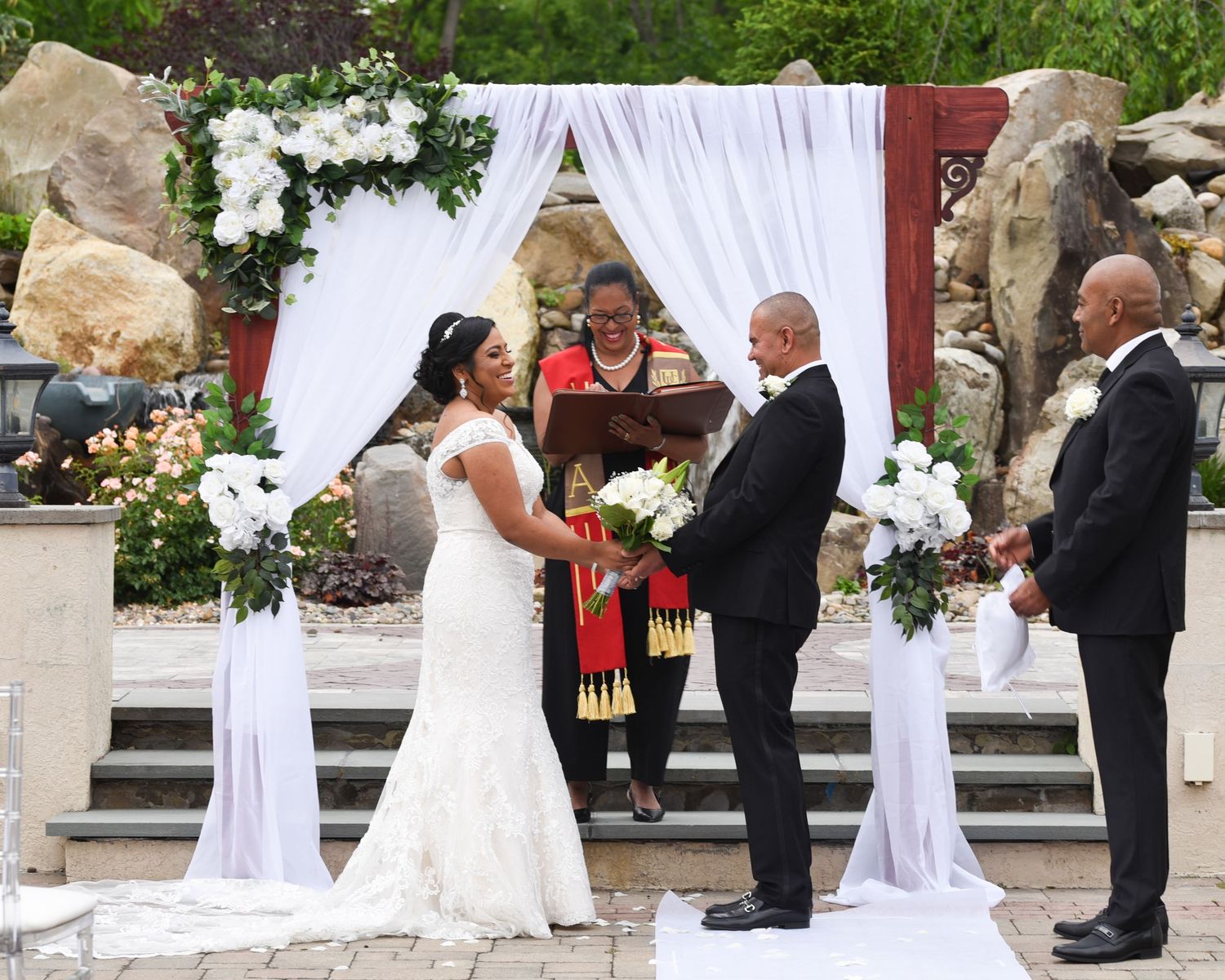 Wedding ceremony taking place under white draped arch with floral decorations and stone backdrop.