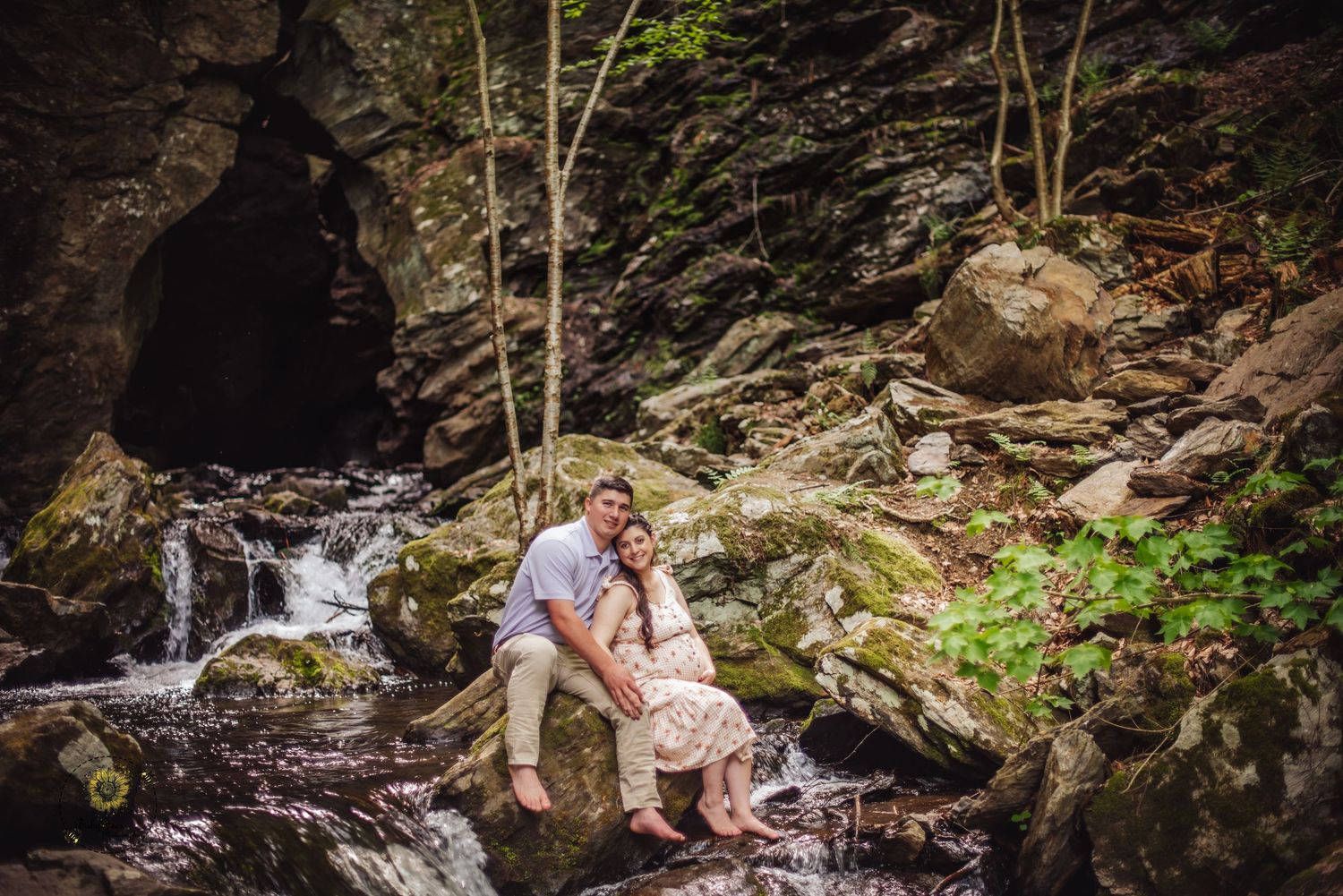 Couple sits together on mossy rocks near a cascading waterfall in a lush forest setting.