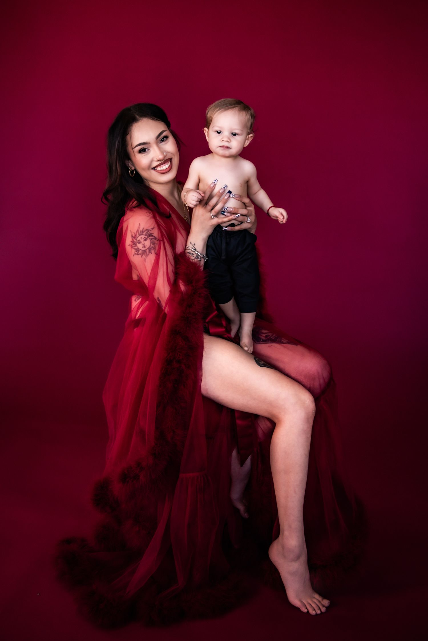Mother and infant photoshoot with flowing red robe creates dramatic silhouettes against burgundy backdrop.