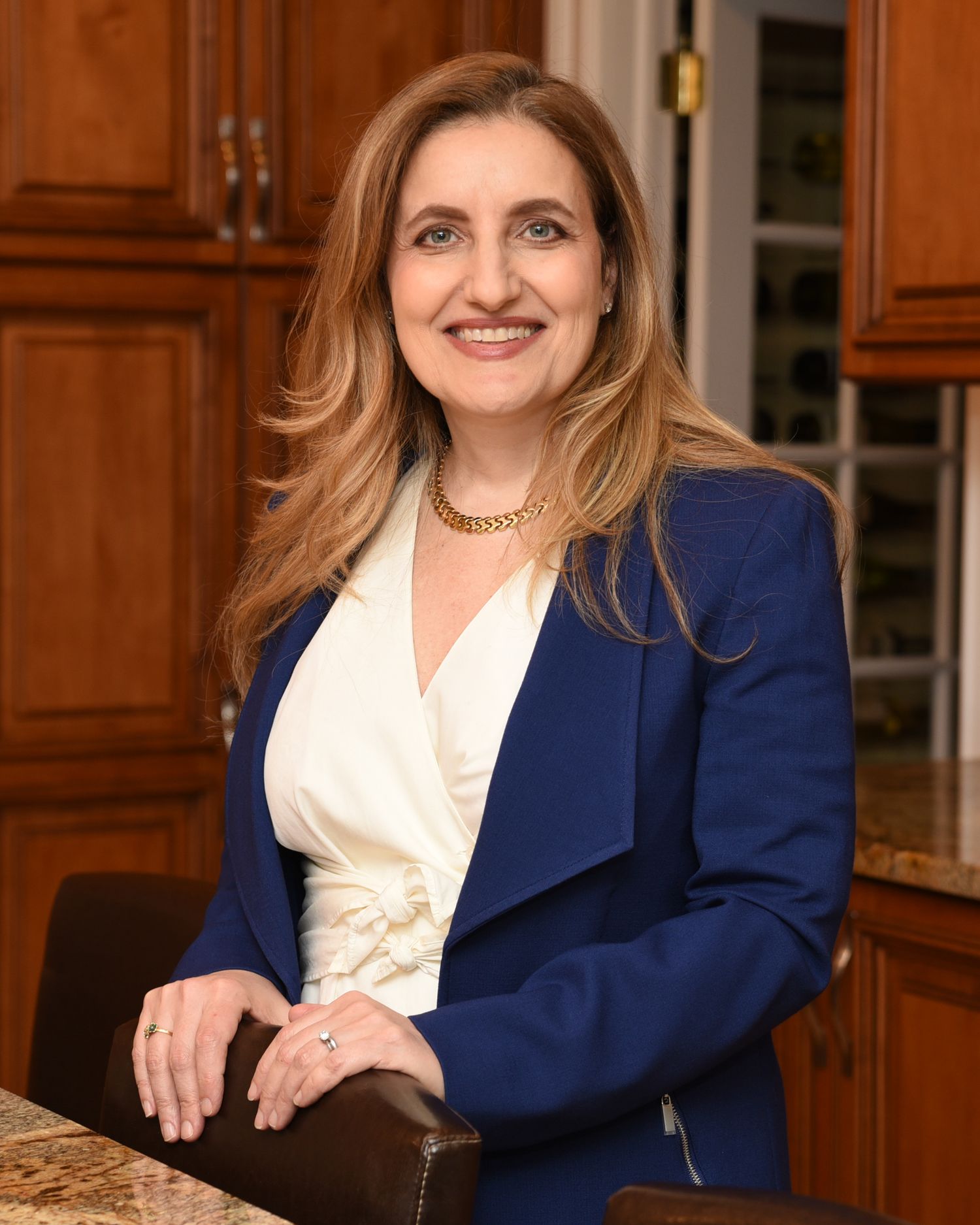 Professional wearing a navy blazer and cream blouse poses at a granite kitchen counter in an elegant setting.