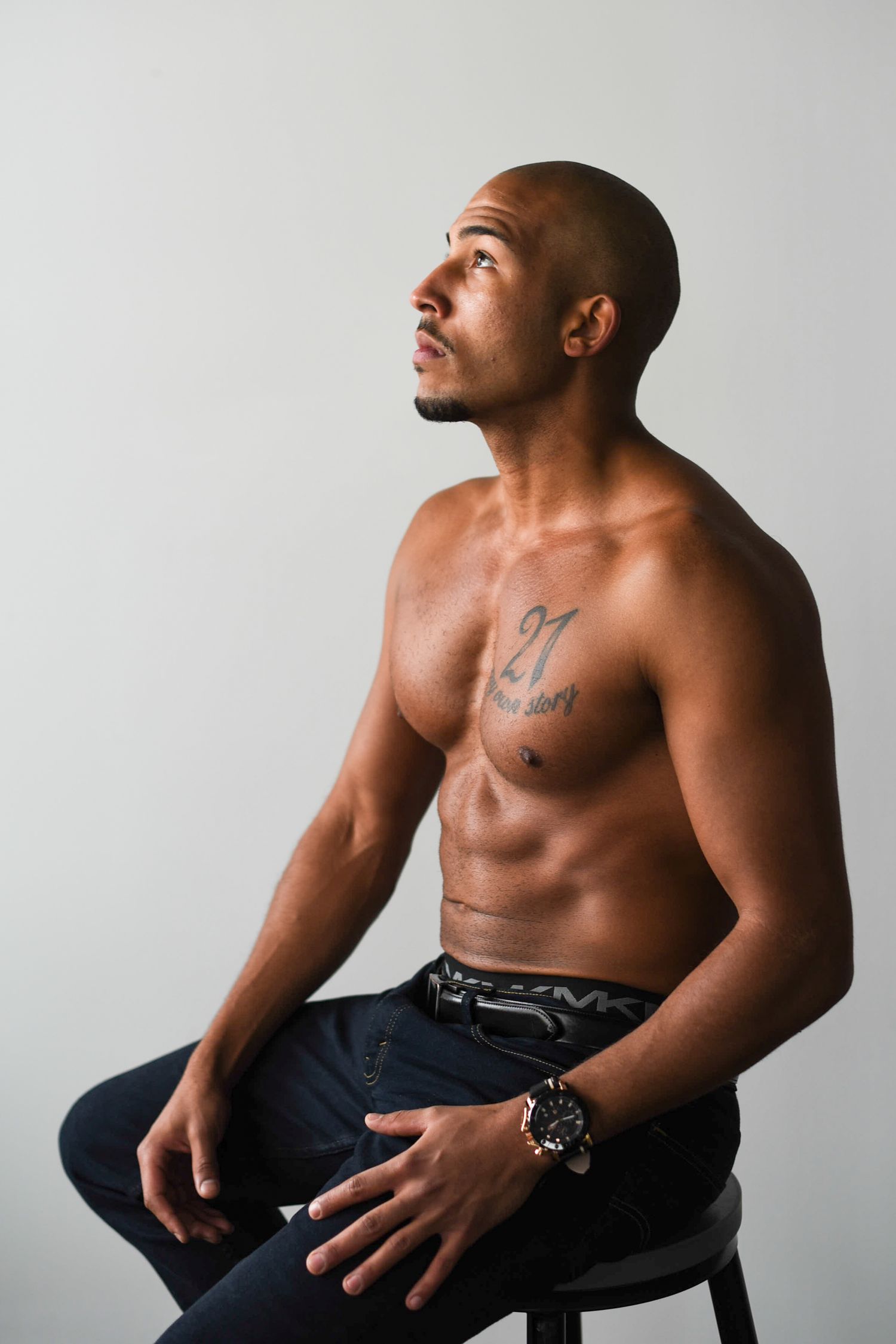 Shirtless muscular man in dark jeans poses against white background in artistic portrait series.
