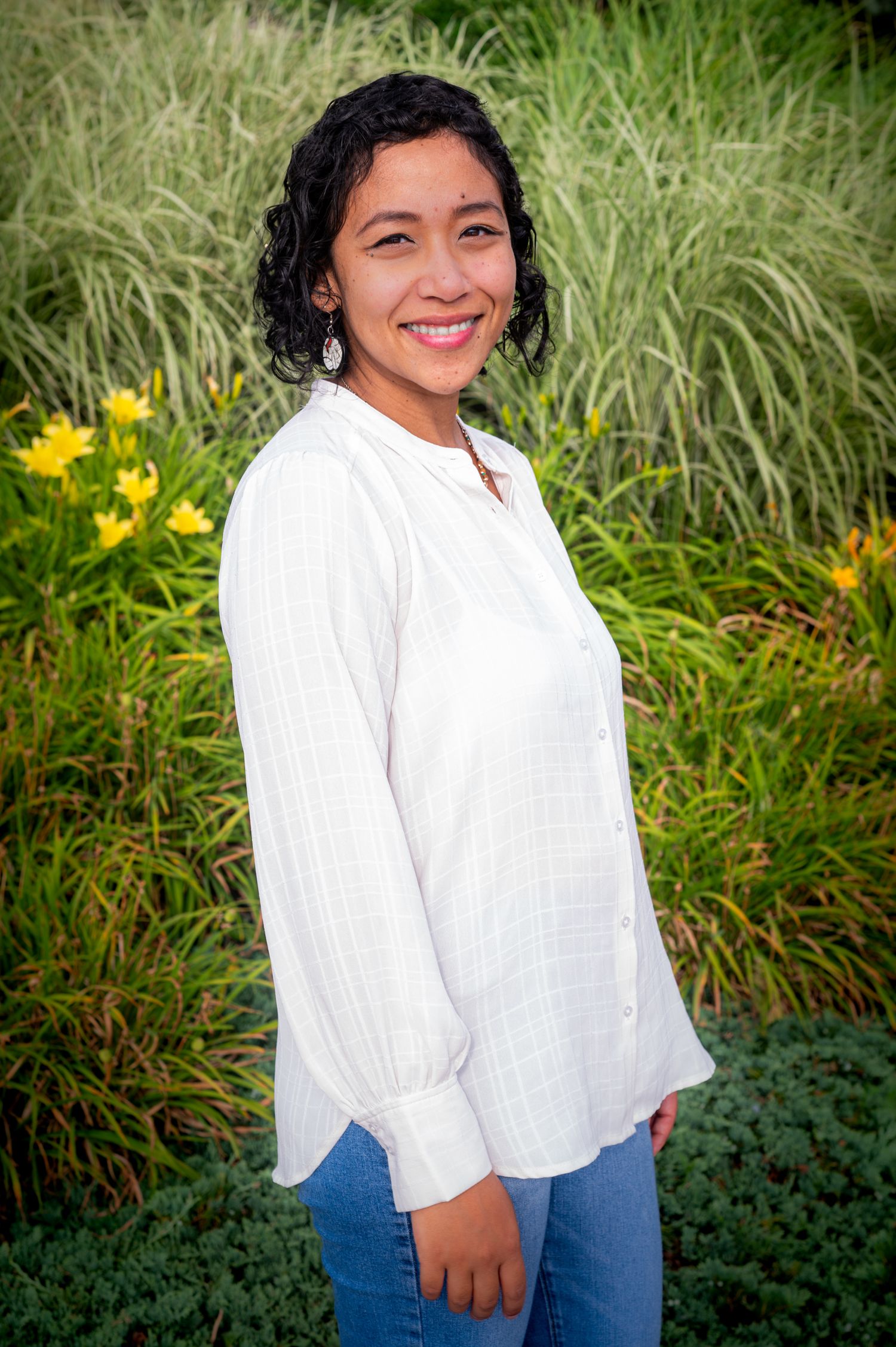 Person in white blouse and jeans poses outdoors against tall grass and yellow flowers in garden setting.