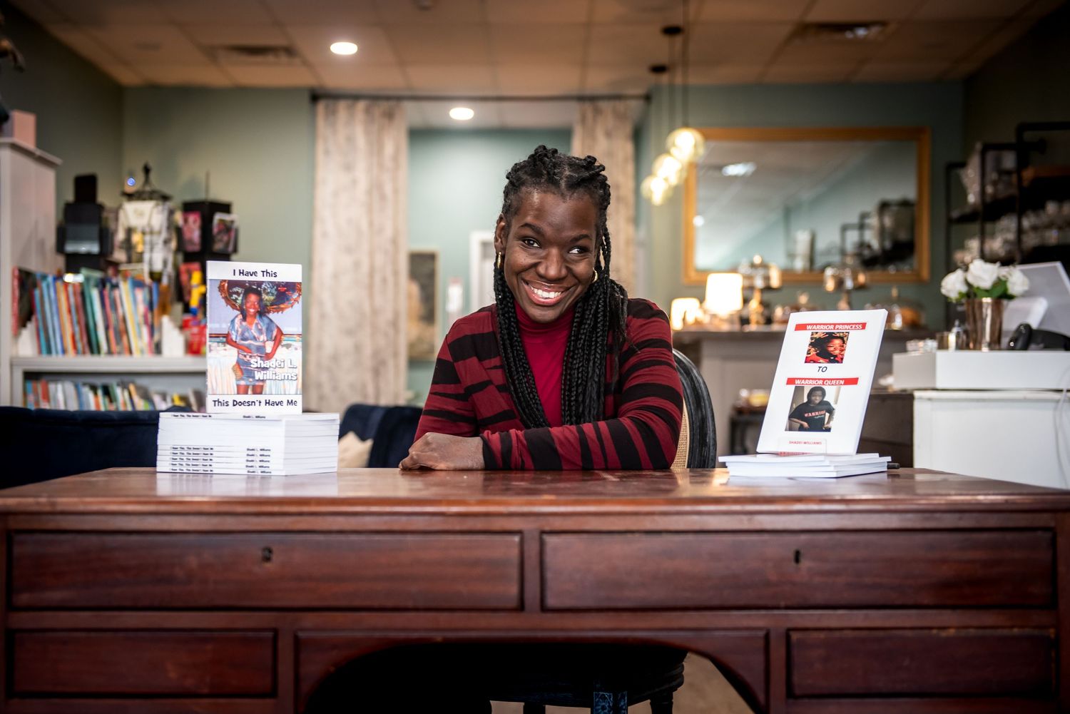 Someone in a red and black striped sweater smiles warmly while sitting at a wooden desk in a library or bookstore.