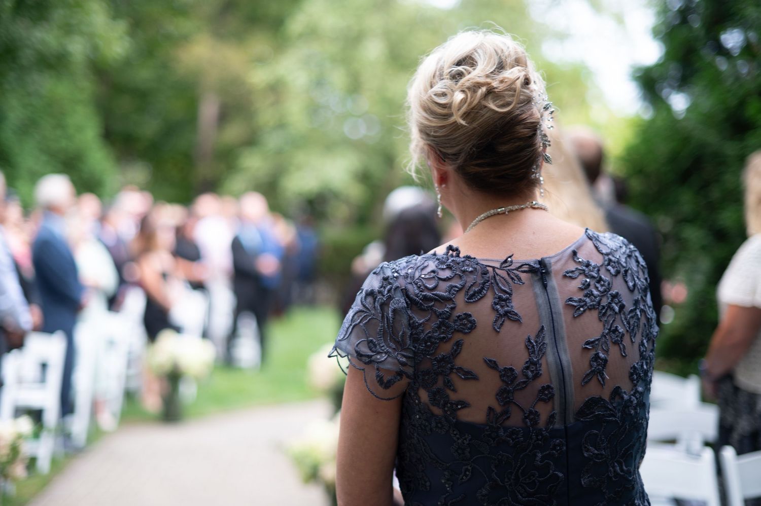 A guest in a navy lace dress watches an outdoor wedding ceremony from behind.